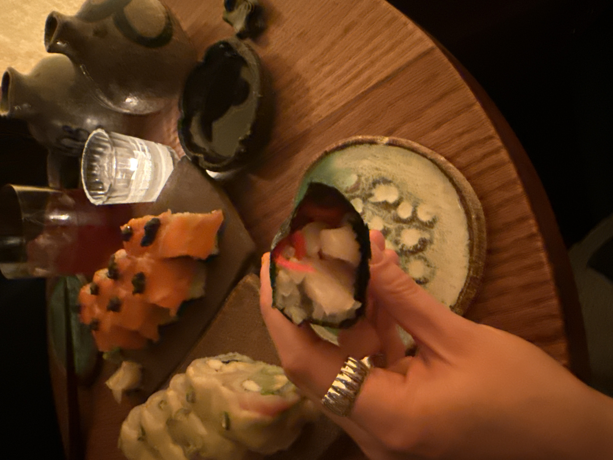 The image shows a hand holding a piece of sushi, with a plate of other sushi rolls on a wooden surface. There are sake bottles and a glass in the background. The location is a restaurant in Mexico City, Mexico.