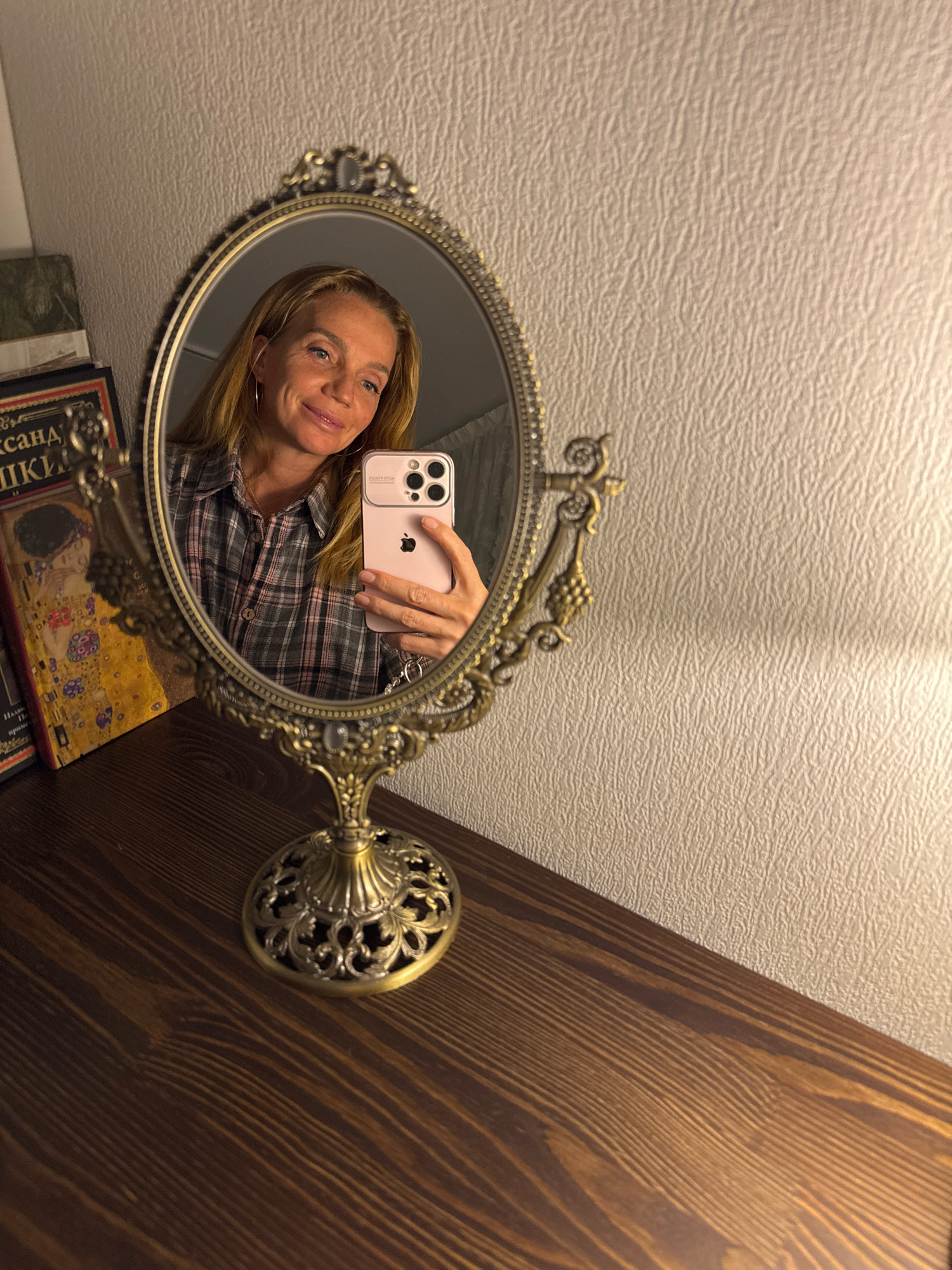 A woman with blonde hair takes a selfie in a decorative oval mirror. She is wearing a grey and pink plaid shirt and silver hoop earrings. She is holding a pink smartphone in her right hand. The mirror is on a dark wooden surface. Behind the mirror, a textured wall is visible. To the left of the mirror are several books, one of which has a gold cover with a painting on it. The image is dimly lit, with warm light illuminating the scene. The location is not identifiable as Veliky Novgorod, Russia.