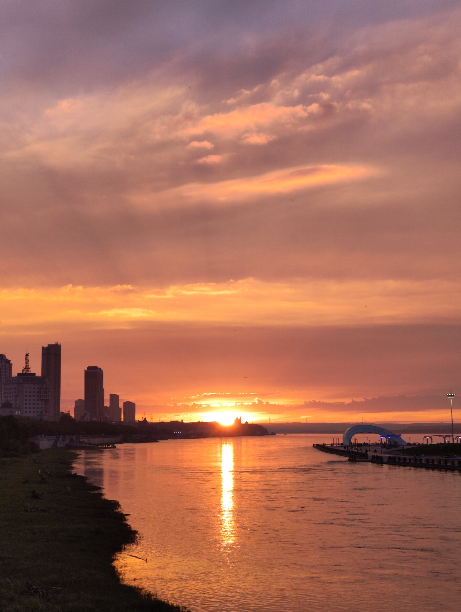 The image captures a vibrant sunset over a city skyline, reflected in a wide river. The sky is a dramatic spectacle of orange, pink, and purple hues, with scattered clouds catching the last rays of the sun. In the foreground, the river flows serenely, its surface shimmering with the reflected colors of the sky. Along the left bank, a grassy embankment is visible, with some scattered debris.
The city skyline is silhouetted against the colorful sky, dominated by tall buildings of varying heights and architectural styles. On the far left, a cluster of buildings forms the initial part of the cityscape. Further to the right, a prominent group of skyscrapers stands tall, with some appearing more modern and angular, while others have a more classic silhouette. There are also some lower structures and a large building with a distinctive antenna on the left side.
On the right side of the image, a modern architectural structure resembling an archway or bridge juts out into the river. This structure is illuminated, and there are people visible walking along it, suggesting it is a public walkway or promenade. A tall lamppost stands near this structure. The distant horizon shows a range of low hills or landmasses, with a few more buildings and structures scattered along the waterfront.
The image is taken from an elevated vantage point, looking across the river towards the city and the sunset. The overall atmosphere is peaceful and awe-inspiring, showcasing the beauty of nature and urban development harmoniously coexisting. The location is identified as 幸福乡, China.
