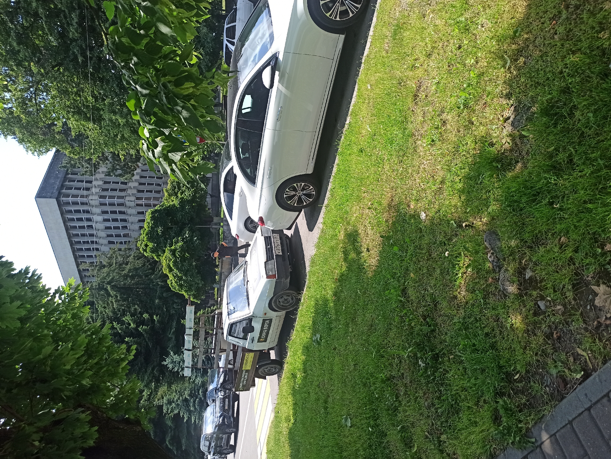 In a scene set in Atabe, Georgia, a row of parked cars lines a grassy roadside next to a sidewalk. The time of day appears to be late morning or early afternoon, indicated by the bright sunlight and distinct shadows cast on the grass. The weather is fair and sunny, with lush green trees flanking the area and a large, modern building visible in the background.
Among the vehicles, a white sedan with a sleek design is prominent in the foreground, parked parallel to the road. Behind it, a white utility truck with a flatbed is parked, displaying visible text on its side. The text appears to be in Cyrillic script and reads "ОКНА БЕРТА" (OKNA BERTA), suggesting a business related to windows. Further down the line, other cars are visible, including a dark-colored SUV. A person dressed in black is seen standing near the utility truck, and another individual in an orange jacket is visible further down the street. The overall setting suggests a typical urban or suburban environment in Georgia during a sunny day.