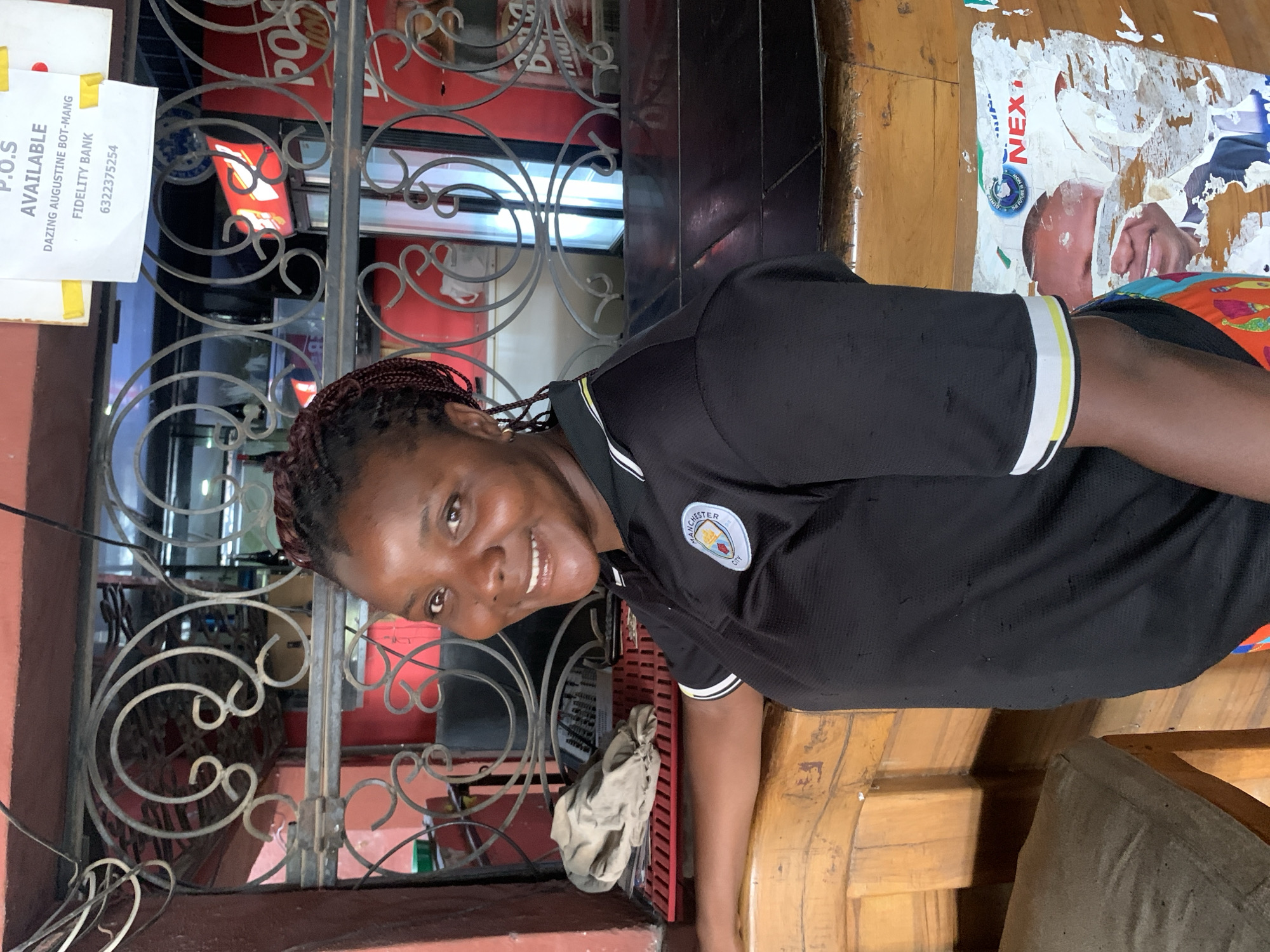 A woman with dark skin and braided hair smiles at the camera. She is wearing a black Manchester City soccer jersey with white and yellow trim on the sleeves and collar. She is standing behind a wooden counter. Behind her is a metal security gate. Through the gate, red signage is visible. A sign on the left of the gate reads, "P.O.S Available," with "Dazing Augustine Bot-Mang" and "Fidelity Bank 6322375254" listed below. The setting is likely a small business in Jos, Nigeria, due to the soccer jersey, P.O.S. (Point of Sale) terminal availability, and general aesthetic. A torn poster of a man is stuck on the counter.