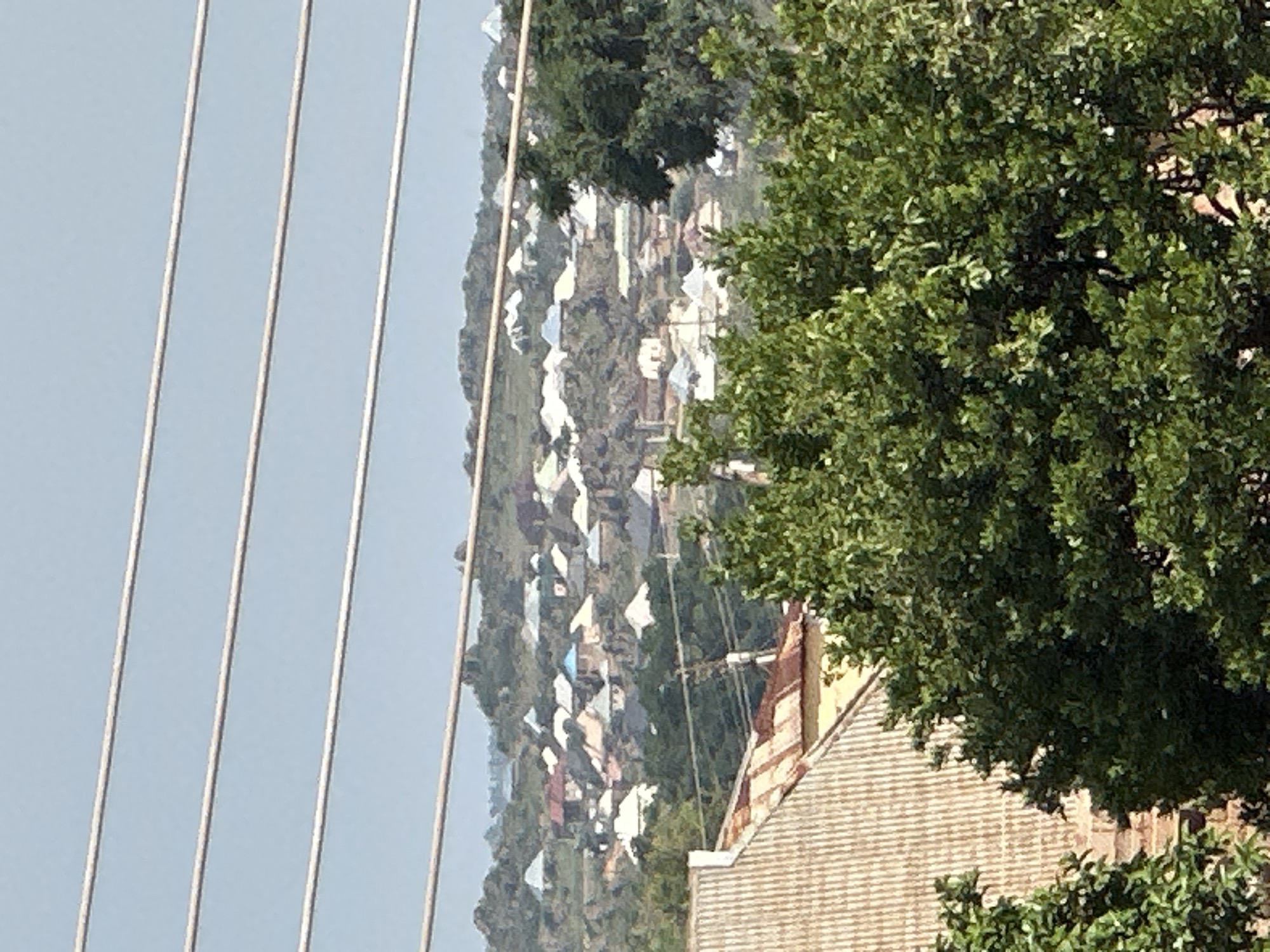 The image displays a view of a hillside settlement in Bukuru, Nigeria. The settlement consists of numerous houses with varied roof colors, including white, blue, and red, spread across the undulating terrain. Green trees dot the landscape, with a large, leafy tree dominating the right side of the foreground, partially obscuring a building with a tiled roof. In the far left of the image, several parallel wires are visible against a clear, light blue sky, suggesting the photograph may have been taken from a high vantage point, possibly from a bridge or a similar elevated structure. The overall scene depicts a residential area nestled within a natural environment, likely during daylight hours given the bright sky and clear visibility. There are no people or discernible activities visible in the image, focusing instead on the architectural and natural elements of the location. No text is visible in the media file.