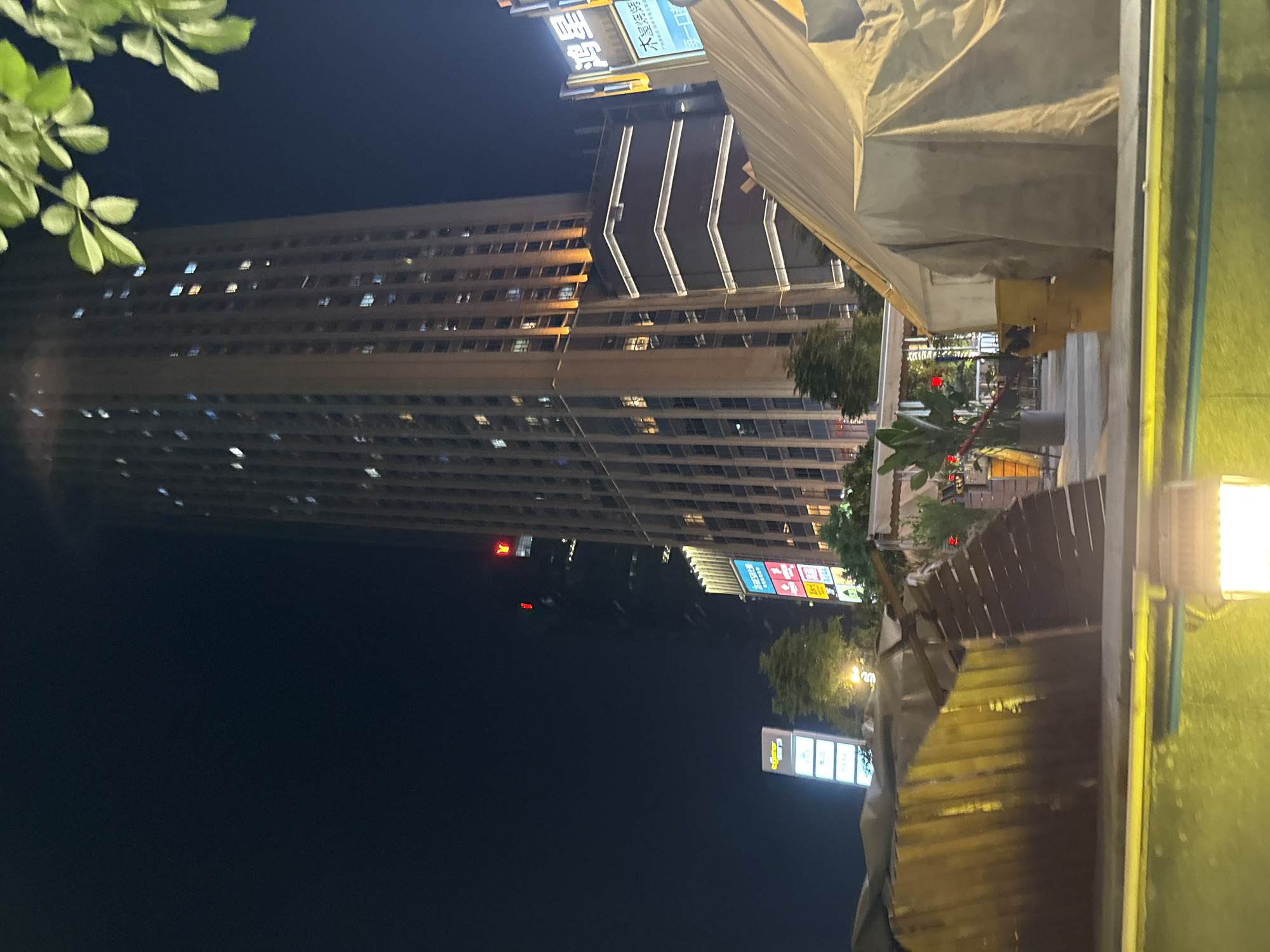 A nighttime scene unfolds in Haizhu District, China, with a focus on urban architecture and signage. Dominating the frame is a tall, modern building characterized by its numerous illuminated windows, suggesting it is inhabited or in use. The building's facade is textured with horizontal lines, and sections of it are highlighted by bright, vertical white lights, creating a striking contrast against the dark sky.
In the foreground, a makeshift structure covered with a large, pale tarp partially obscures the view of another building and some vegetation. This structure appears to be temporary, possibly a stall or construction covering. To the right, a bright outdoor light illuminates a green surface, possibly a wall or a tarp, casting a warm glow.
Several signs are visible, adding to the urban feel of the location. One sign, predominantly blue and white with some red elements, features text in what appears to be Chinese characters. Another, further down, is a colorful sign with more Chinese characters. A small, illuminated red sign is also visible on one of the buildings.
The time of day is clearly night, indicated by the dark sky and the illuminated windows and signs. The weather is not explicitly discernible, but the overall atmosphere suggests a typical urban night in a developed area. The scene conveys a sense of everyday urban life at night, with the architecture and commercial signage providing specific clues to its location and nature. No people are explicitly visible, and the focus is on the static elements of the environment.