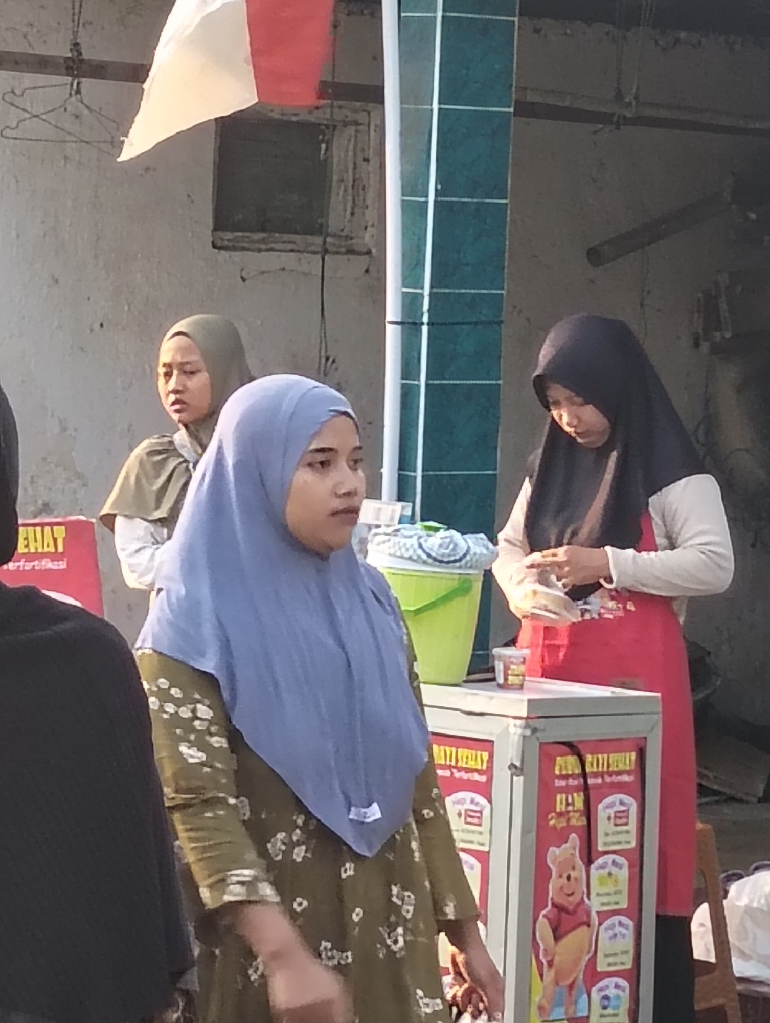 The image depicts an outdoor scene in Banjarwati, Indonesia, likely a market or roadside stall. Three women are visible. In the foreground, a woman wearing a light blue hijab and a green floral patterned dress is looking to her right. To her left, another woman in a black hijab and a cream-colored shirt with a red apron is engaged with a clear plastic package in her hands. Behind these two women and slightly to the left, a third woman wears a light green hijab and a dark top. A red and white flag is visible in the upper left corner. A green bucket and a white cooler with various labels, including an image of Winnie the Pooh, are present in the scene. The background shows a textured wall and some overhead structures. The lighting suggests it is daytime.