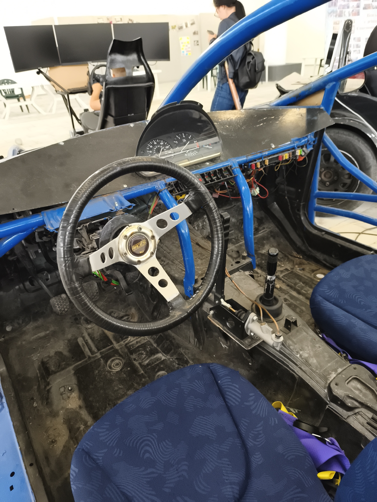 The image shows the interior of a car-like vehicle with a blue roll cage. The dashboard is black and contains a speedometer. A steering wheel with a "RACES" branded center is visible. A gear stick and some mechanical components are near the seats, which are blue with patterned fabric. The vehicle interior appears unfinished and dirty. In the background, there are people, office furniture, and computer monitors. Based on image metadata, the location is Targovishte, Bulgaria.