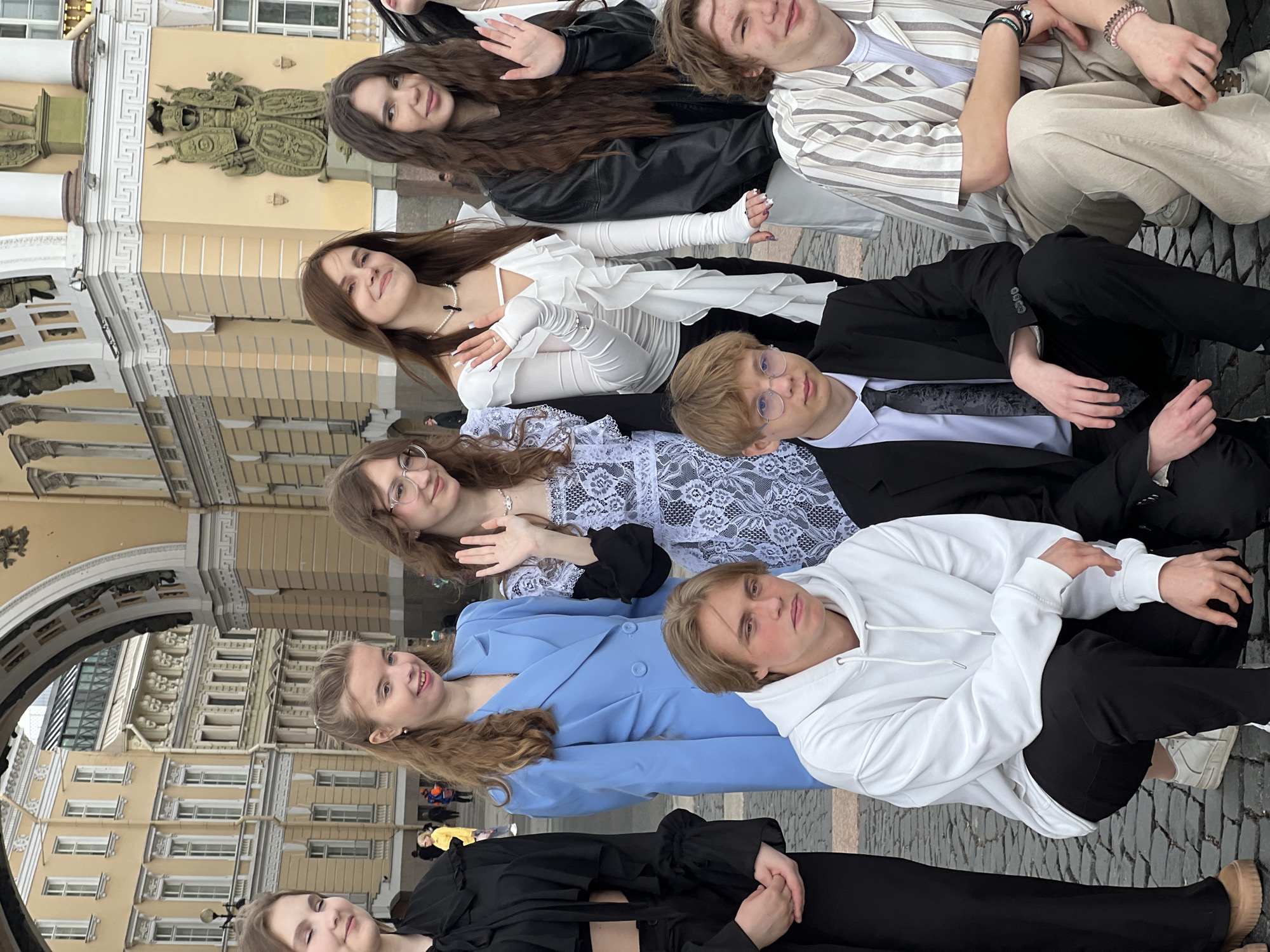 A horizontal, eye-level photograph shows a group of young people posing together in Saint Petersburg, Russia, in front of a portion of the General Staff Building on Palace Square. The group consists of seven individuals. From left to right, the first person is in a long black dress. Next to her is a person in a blue blazer. The person next to them is in a white hoodie. To the right of this person is one in a white lace dress with a black trim. Another person is positioned in the middle wearing glasses and a black suit. The person next to them is in a white dress, and the final person is in a black leather jacket and a beige striped jacket. In the background, there is a portion of the General Staff Building with its distinctive architecture, including windows, columns, and decorative elements. The scene is outdoors, and the lighting suggests a daytime setting. The ground is paved with stone. The composition captures a casual moment with the group, likely a commemorative photograph.