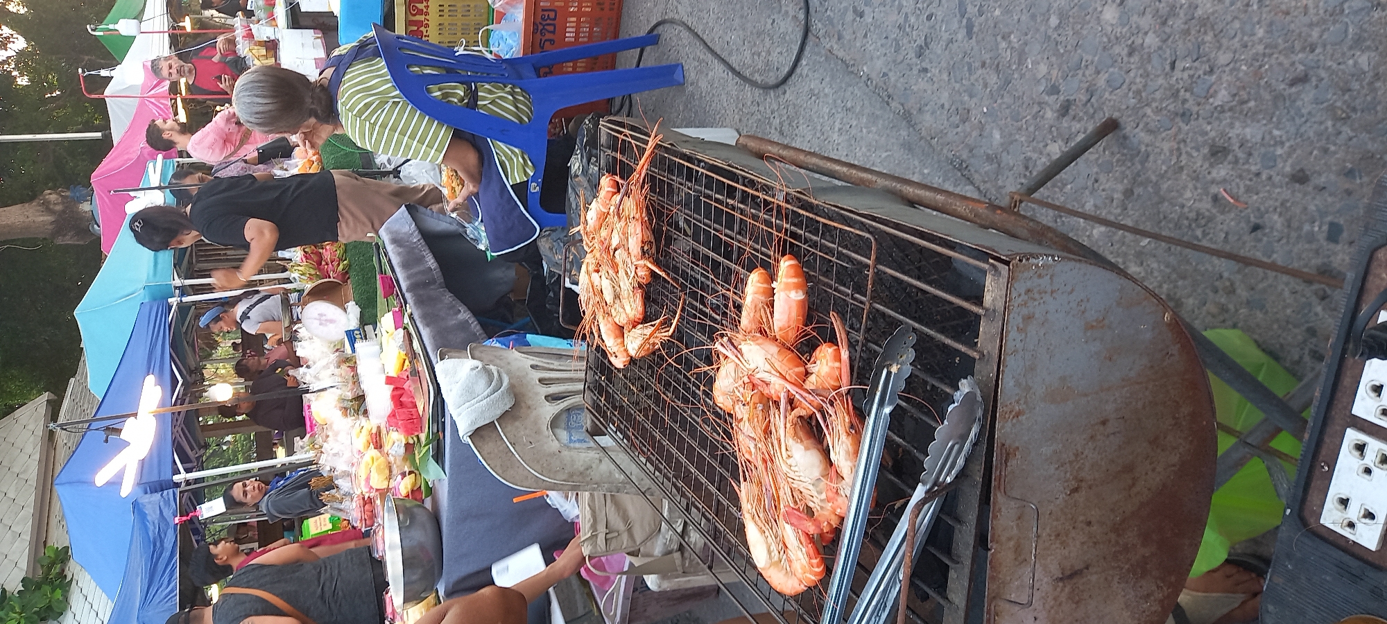 The scene depicts a street food stall in Thong Sala, Thailand. A vendor in a green-striped shirt is attending to customers near a grill laden with large prawns. The prawns are in various stages of cooking. Next to the grill, there is a pair of metal tongs. Customers browse other food items at stalls under tents. The area appears to be a busy market or street fair.