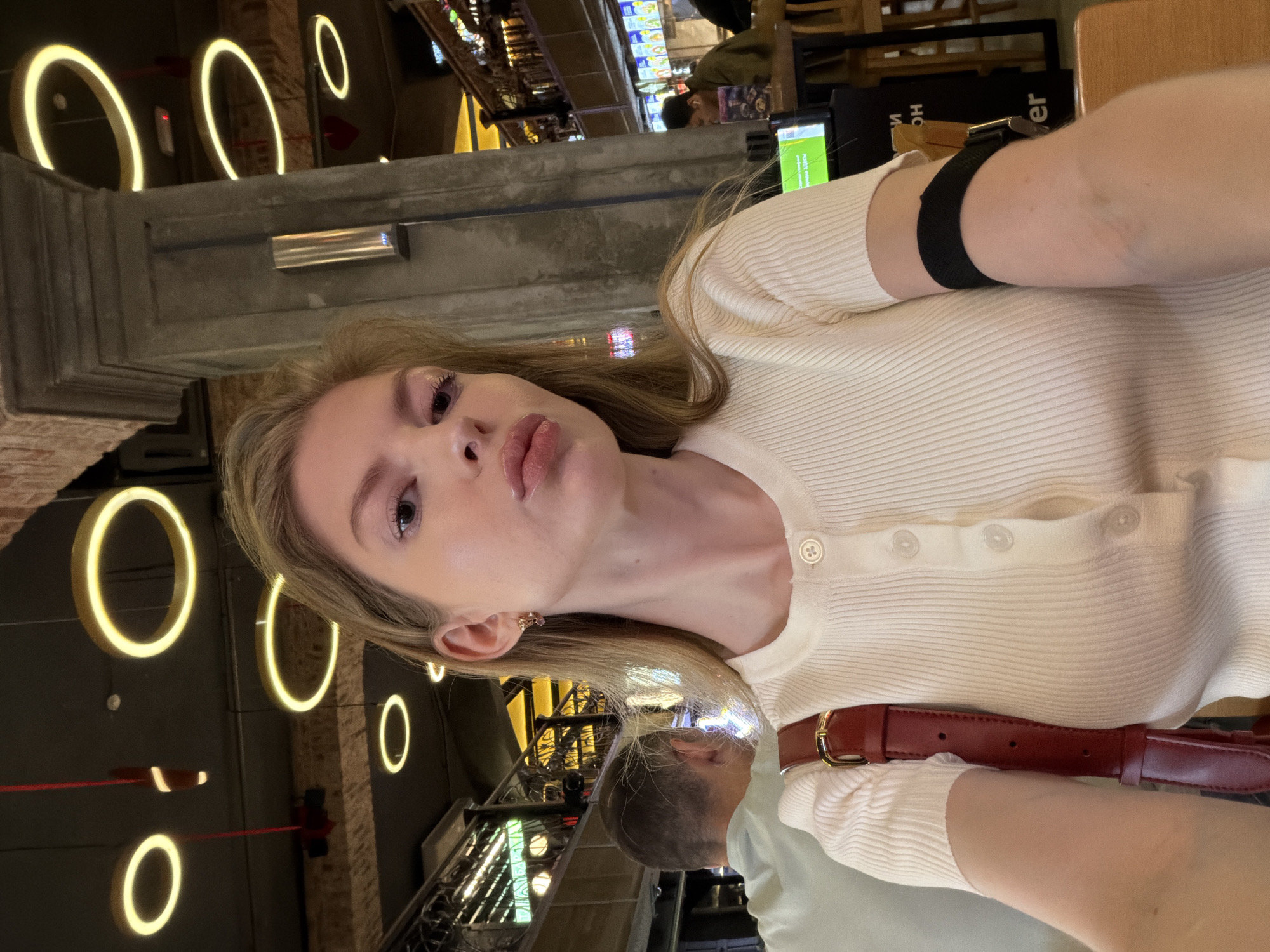 A selfie shows a fair-skinned woman with light hair in an indoor setting with a high ceiling. She wears a light-colored, button-down top and a black wristband. A red belt with a gold buckle is visible. In the background, there are several halo-shaped lights hanging from the ceiling. A man is partially visible near the woman. There appears to be a bar or restaurant in the background, with shelves of bottles and other patrons. The scene is in Moscow, Russia.