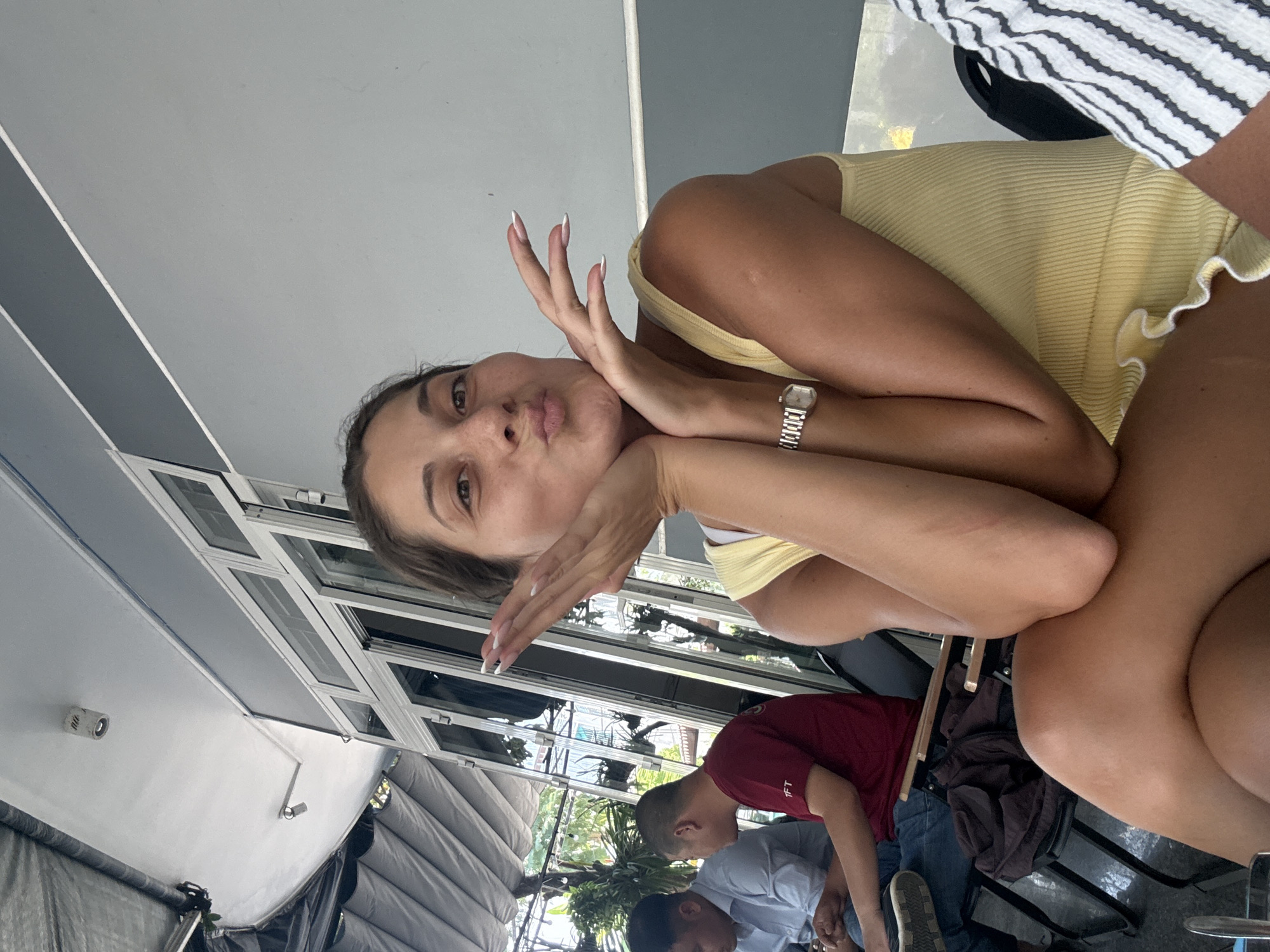 A woman in a yellow top with a silver watch is posing for a photo. Two men are in the background. The location may be a restaurant in Đà Nẵng, Vietnam based on the plants visible through the windows and general style of the building.