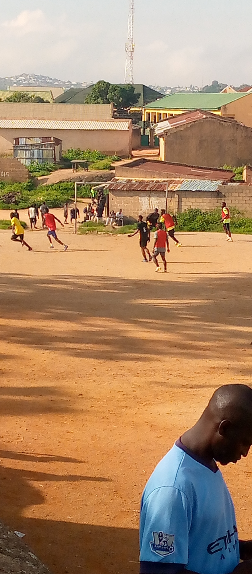 A group of young men are playing a soccer match on a dusty, outdoor field. The game appears to be informal, with players wearing a mix of athletic attire and casual clothing. In the background, there are buildings with corrugated iron roofs, some residential and others appearing to be small businesses. A cell tower is visible in the distance. The scene suggests a typical day in an urban or semi-urban environment in Jos, Nigeria.