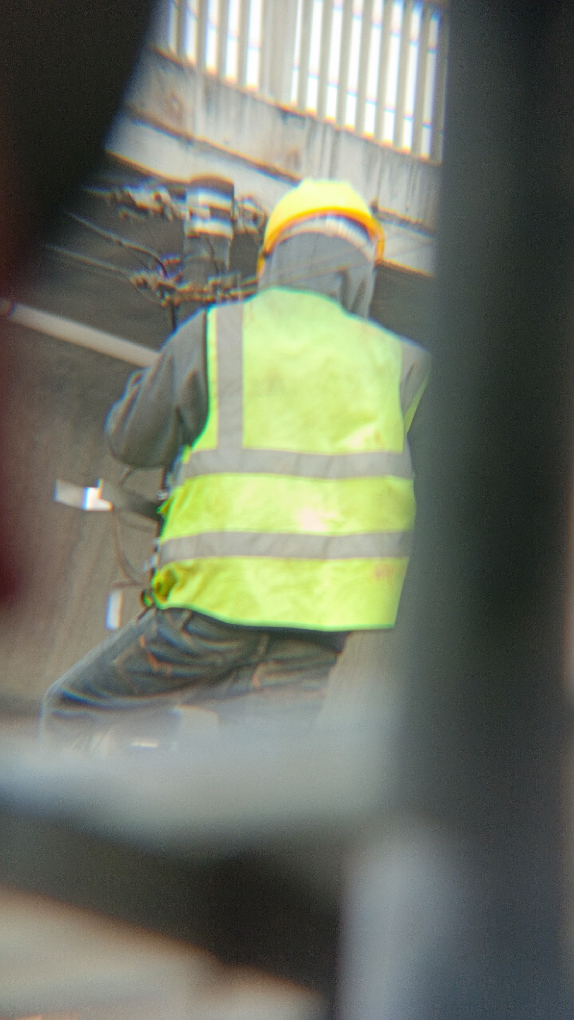 The image shows a construction worker wearing a yellow hard hat and a neon yellow safety vest over a gray hooded jacket. The worker is also wearing blue jeans. The worker appears to be in the process of working, as indicated by their attire and posture. The background suggests an outdoor urban environment, possibly a construction site, with some infrastructure visible, including what looks like a bridge or elevated walkway with vertical elements, and possibly some piping or electrical conduits. The overall image has a blurred and somewhat distorted quality, with chromatic aberration visible, which might be due to the lens used or the photographic conditions. The specific location in Margajaya, Indonesia, is not visually identifiable within the image itself without external context.