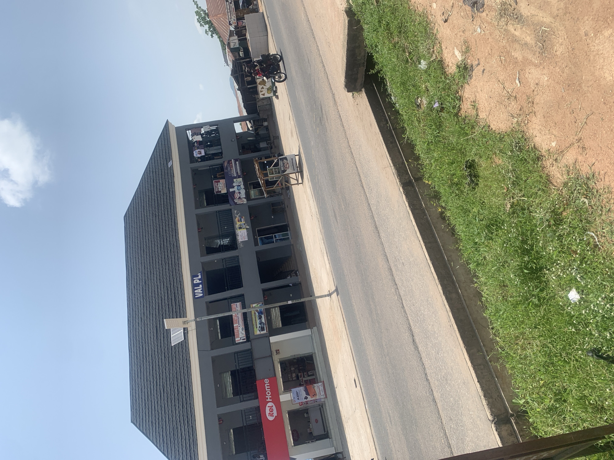 The image captures a street scene in Jalingo, Nigeria, focusing on a modern two-story building with a dark tiled roof. The building, labeled "VAL PL." at the top, features a grey facade with multiple shops on the ground floor. One prominent shop displays a red "itel Home" sign. The upper floor has several balconies or openings with railings. A solar-powered street light stands in front of the building. A paved road runs alongside the building, with a drainage ditch and grassy area on the right side of the image. In the foreground, a motorcycle with two riders is traveling on the road. The sky is clear with a few scattered clouds. The surrounding environment suggests an urban setting, possibly a commercial area.