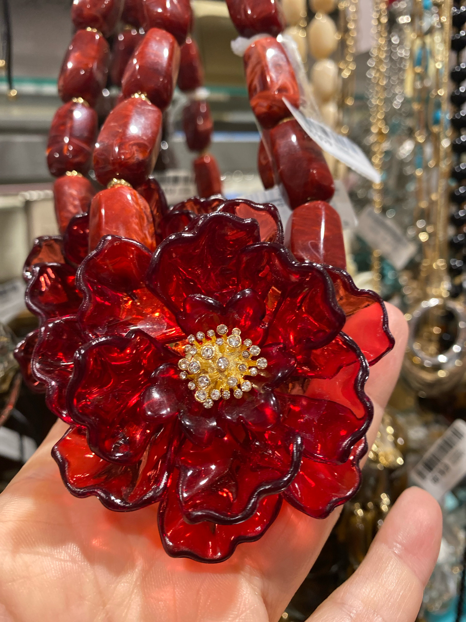 A close-up, eye-level shot features a person's hand holding a large, ornate, red, translucent floral pendant. The pendant is intricate, with layered petals and a central embellishment of small, clear rhinestones set in gold. Behind the pendant, the blurred background reveals a display of various necklaces and jewelry pieces, suggesting a retail environment. Some of the jewelry in the background appears to be gold, black, and possibly turquoise. The lighting is bright, with a shallow depth of field, making the pendant the primary focus. The scene is indoors. The image does not provide any information to determine its location in Vienna, Austria.
