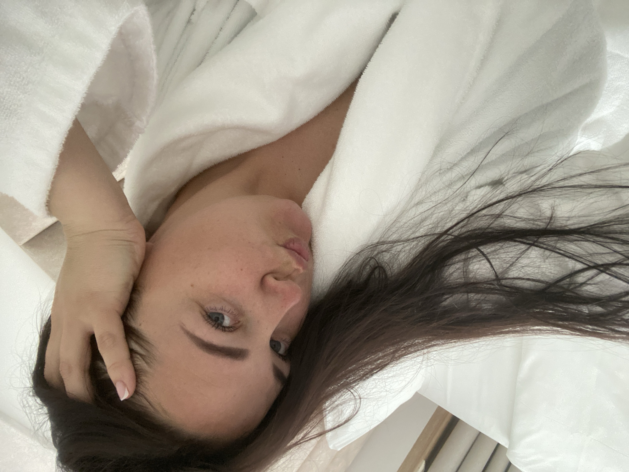 A woman is lying on a white surface, presumably a bed, wearing a white bathrobe. She has dark hair and fair skin. Her right hand is resting on her head. The environment appears to be indoors, possibly a hotel room or home setting. There is no information provided that indicates that this image was taken in the city of Sochi, Russia.