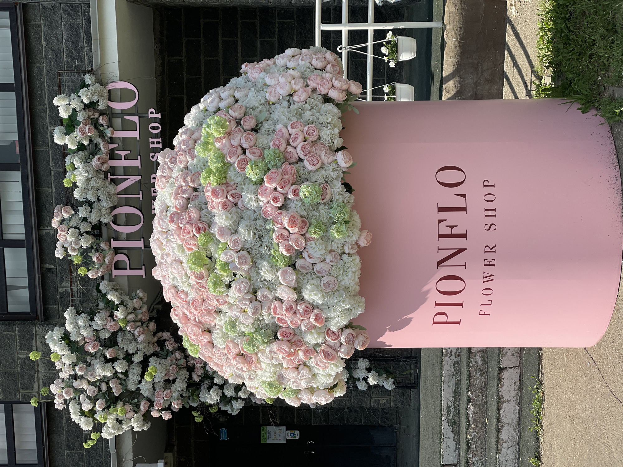 The image shows a large, pink, cylindrical planter filled with an abundance of artificial flowers. The planter is situated outside a building with a dark brick facade. Above the planter, a pink sign reads "PIONFLO FLOWER SHOP". Pink and white flowers cascade down the side of the building, adorning a black wrought-iron gate. White hanging baskets with small white flowers are suspended from a white railing to the right of the planter. The scene appears to be a storefront decorated for a special occasion or as a permanent aesthetic feature. The location is identified as Kazan, Russia.