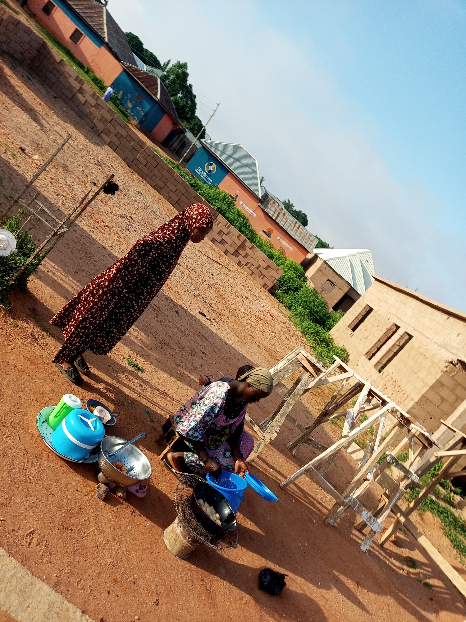 In Tempure, Nigeria, two women are engaged in activities outdoors on a dirt surface, with buildings in the background. The woman on the left, wearing a dark patterned dress and headscarf, stands facing right. In front of her are several plastic containers, including a blue one with a white lid, a green container, and a silver bowl. To the right, a second woman, wearing a colorful patterned dress and headscarf, is seated and looking downwards, appearing to be cooking over a small charcoal stove. Next to her is a blue plastic bucket and a blue ladle. The environment shows construction materials, including bricks and wooden scaffolding, indicating ongoing building work. The sky is clear and blue.