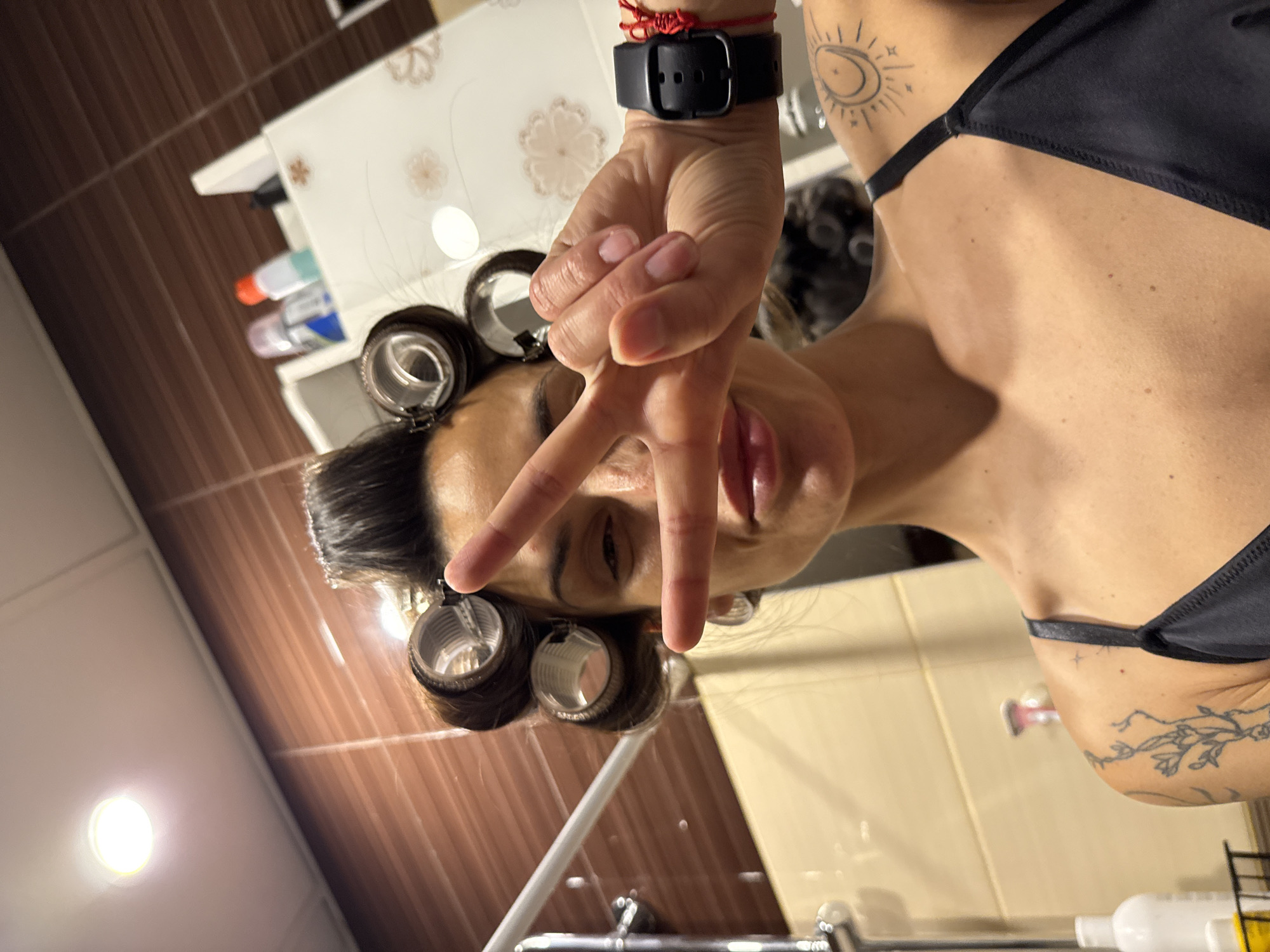 A medium shot depicts a woman with hair rollers posing in what appears to be a bathroom. Her face is visible, with her hand in front making a peace sign. She wears a black bra and a smartwatch with a black band and a red bracelet. Her neck and upper chest are visible, revealing a tattoo near her right shoulder. The background includes a sink, a cabinet with toiletries, and beige tiles on the wall. Based on the details, there is no visible information to confirm the location in the city of Pleven, Bulgaria.