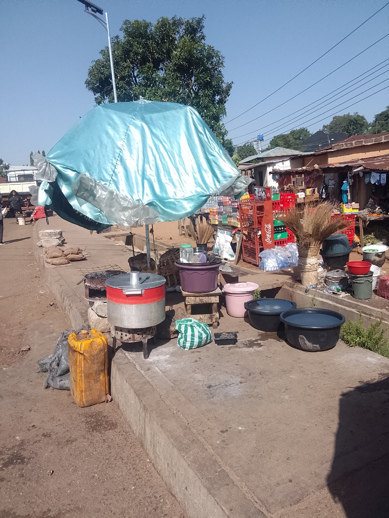 The image depicts a street scene in an unidentified city. A blue-silver umbrella provides shade to a cooking area on a concrete sidewalk. The cooking area consists of a red and silver metal pot on a stand above a small charcoal brazier, with additional pots and basins nearby. A yellow jerrycan is positioned to the side. In the background, there's a small roadside shop stocked with beverages, including Coca-Cola. Trees and other buildings are visible in the background under a clear sky.