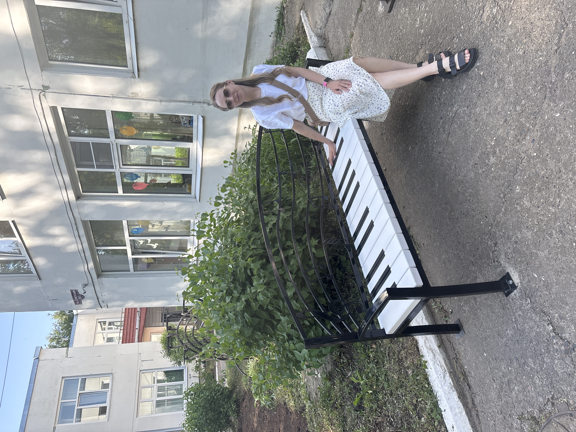 A woman sits on a public bench designed to resemble a piano keyboard in the city of Chaikovsky, Russia. The bench is black and white, and sits next to a leafy green bush. The woman has long hair, is wearing sunglasses, a white top, and a floral print skirt. Behind her, a multistory building with windows can be seen. The ground is paved with gray asphalt.