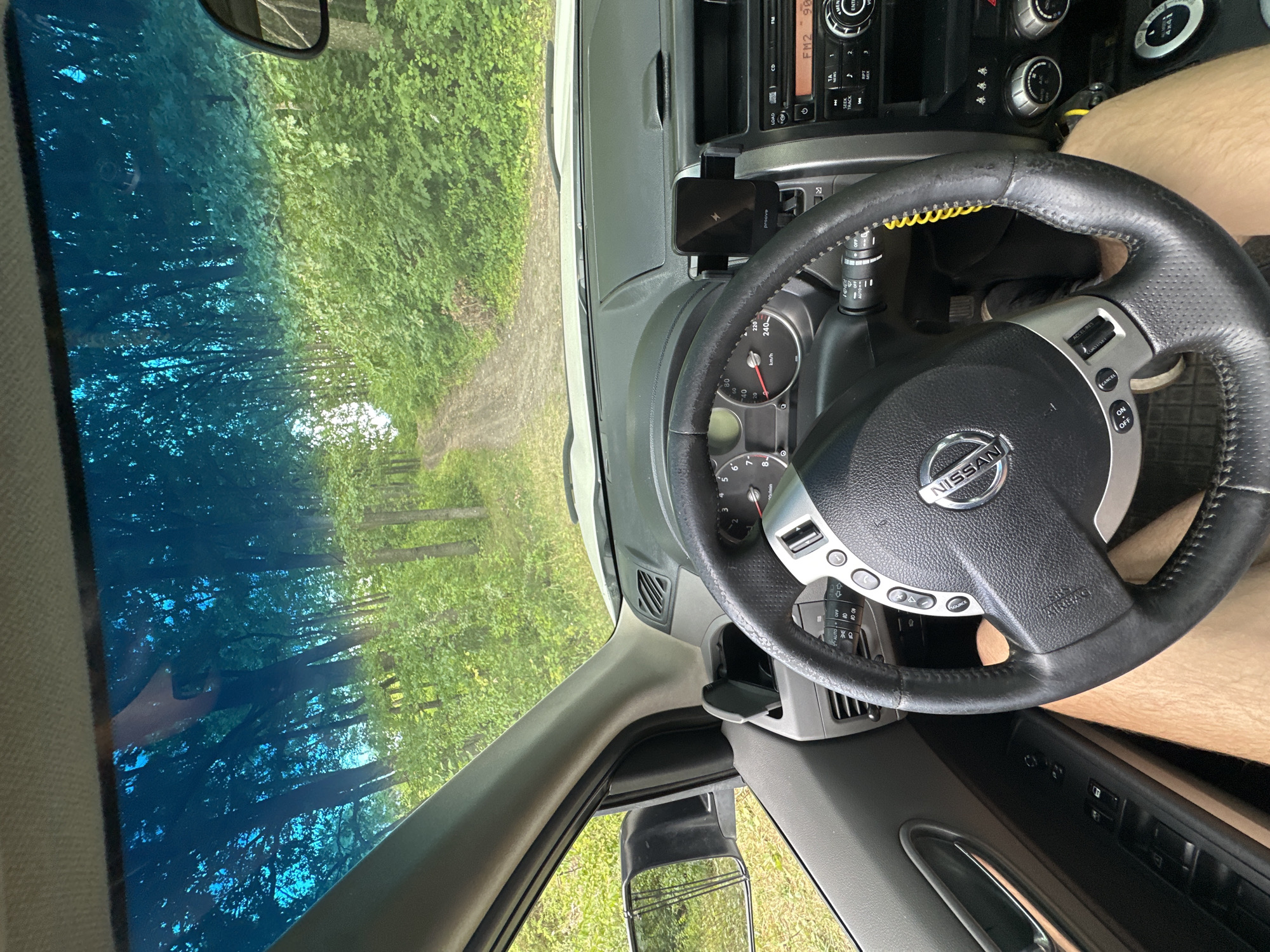 The image shows the interior of a Nissan vehicle, specifically the dashboard and steering wheel, with a view through the windshield. The vehicle appears to be on an unpaved road surrounded by dense green foliage, characteristic of a rural or forested area. The road itself is dirt and appears to be a path leading through a wooded area. The sky visible through the windshield has a bluish tint.
Inside the car, the steering wheel is prominent, with a Nissan logo at its center. Various controls and dashboard elements are visible, including the instrument cluster, air vents, and a car stereo. A phone mount with a wireless charger is attached to the dashboard. The driver's legs are visible in the lower part of the frame, suggesting the photo was taken from the driver's perspective. The side mirror is also visible on the left, reflecting the surrounding greenery.
The image was taken in Makiv, Ukraine.