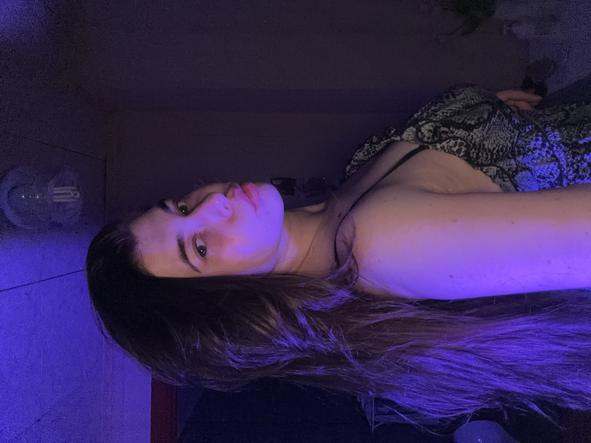 A young woman with long brown hair lies on a dark surface, illuminated by a blue light. She is wearing a snake-print dress. The location is likely not Мещёры, Russia, as the image provides no information to support that claim. There is a light fixture visible in the background. The woman appears to be posing for the image.