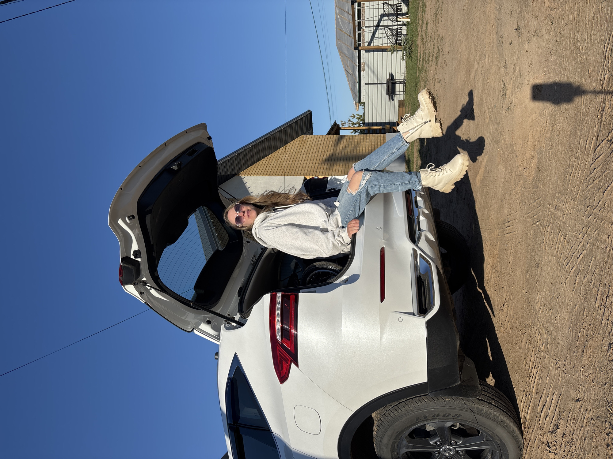 A young woman sits in the open hatchback of a white SUV. She is wearing sunglasses, a grey sweatshirt, ripped jeans, and beige combat boots. She is leaning back, with her legs extended out of the vehicle. The car is parked on a dirt surface, with a house visible in the background. The sky is bright blue. The location appears to be in a rural or suburban area, as indicated by the dirt ground and the style of the house in the background. There is no visual confirmation that the location is Chaikovsky, Russia.