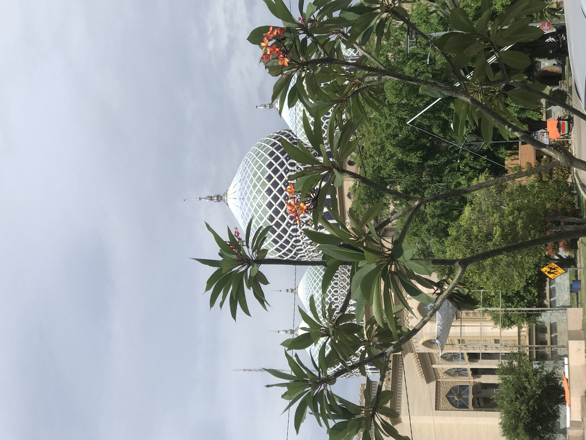 The image shows a mosque in Lhokseumawe, Indonesia. Two domes with a diamond lattice pattern are visible. A plumeria tree with orange flowers frames the mosque. There is also an adjacent building next to the mosque with arch windows. The sky is cloudy. In the background, green trees and a yellow caution road sign can be seen.