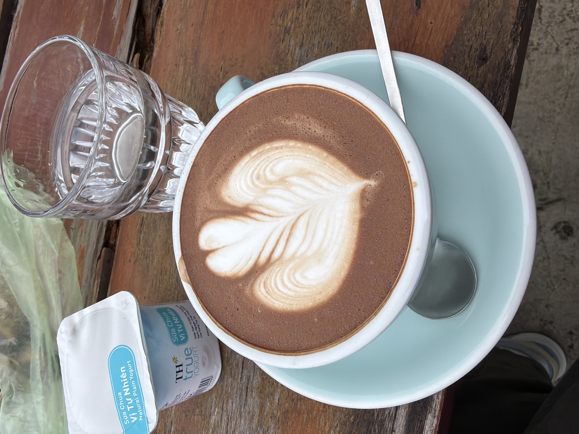 The image depicts a close-up, top-down view of a table setting at an outdoor cafe. The table is made of weathered, dark brown wooden planks. In the foreground, a light blue saucer holds a white mug filled with a frothy, chocolate-colored beverage. The beverage has latte art in the center, resembling two symmetrical hearts with feathered accents. A silver spoon rests on the saucer to the right of the mug. Behind the mug, to the left, is a small, white plastic container of "TH true YOGURT" labeled in Vietnamese and English as "Sữa Chua Vị Tự Nhiên Natural Plain Yogurt". To the right of the yogurt, a clear glass filled with water sits on the table. The background shows a blurred green plastic material, likely a bag or covering, and more of the wooden table. The overall impression is of a simple, casual breakfast or snack. The scene is consistent with the ambiance of many cafes in Da Lat, Vietnam, known for their natural settings and local produce.