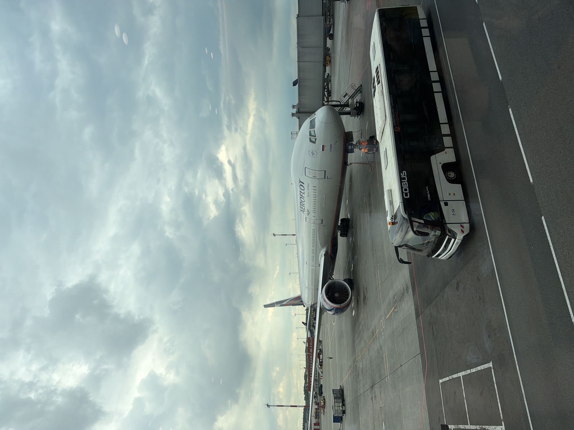 An aerial view shows a cloudy sky over an airport tarmac in Khimki, Russia. A large Aeroflot airplane, identified by its "AEROFLOT" branding and Russian flag livery, is parked at a gate. A white COBUS airport bus is positioned in the foreground, partially obscuring the tarmac. Two individuals in orange vests are visible near the airplane's boarding stairs, suggesting ongoing ground operations. The tarmac is marked with yellow and white lines, indicating parking and movement areas. Other airport infrastructure, such as jet bridges and utility vehicles, are visible in the background. The overall scene depicts a typical airport environment under overcast conditions.