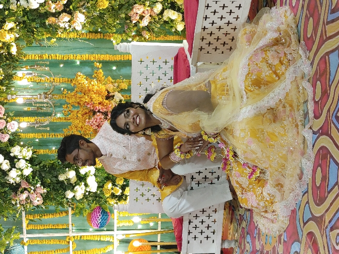 The image captures a posed scene featuring two individuals, one male and one female, at a celebratory event in Dhaka, Bangladesh. The male, positioned on the left side of the frame, is seated on a white, ornate wooden bench with a red cushion. He wears a yellow long-sleeved garment, white trousers, and a white, embroidered Nehru jacket, accessorized with glasses and a beard. He is smiling and looking towards the left. The female, positioned to the right and in front of the male, is seated on the floor. She wears an elaborately embroidered yellow lehenga, a matching choli, and a sheer yellow dupatta, with flower garlands and bangles on her wrists. Her dark, curly hair is styled, and she is smiling while looking towards the right, with her hands clasped in front of her. The background features a decorated stage or backdrop adorned with vertical strings of yellow marigold flowers, green leafy garlands, and arrangements of white and light pink roses. A silver-colored chandelier with multiple lights hangs centrally, and a multi-colored honeycomb lantern is visible on the far left. The floor is covered with a patterned rug in shades of red, blue, and gold.