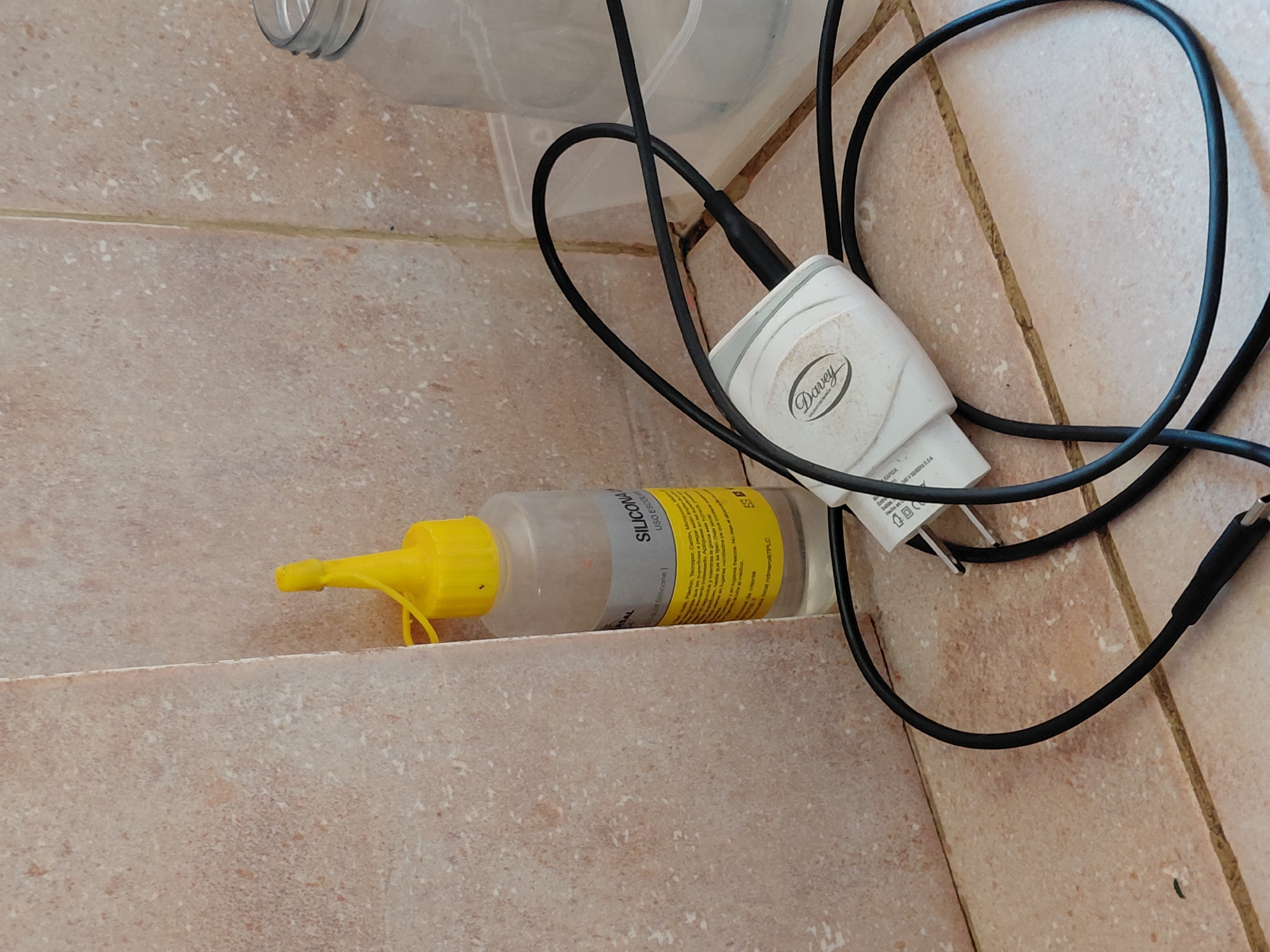 The image depicts an indoor scene with several objects resting on a tiled surface. The specific location in the city of Santa Ana, Venezuela, cannot be determined from the visual information provided.
The environment consists of light beige or light brown tiles, possibly ceramic, covering both a floor and a wall, with a visible corner where they meet. The tiles have a granular or speckled texture. Grout lines separating the tiles are visible, with some showing discoloration or accumulation of dirt.
On the tiled floor, positioned near the wall, is a translucent plastic bottle with a bright yellow, conical dispenser cap. A label on the bottle is partially visible, indicating "SILICONA LIQUIDA USO ESCOLAR" (Liquid Silicone School Use). The bottle contains a clear liquid and appears partially full.
Adjacent to the bottle, further into the foreground, is a white power adapter (charger) for an electronic device. The brand name "Davey" is embossed on its surface. A black USB cable is plugged into the adapter, and a portion of the coiled cable rests on the tiled surface. Both the adapter and cable show signs of light dust or dirt accumulation.
In the background, to the right, an empty, clear plastic jar or wide-mouthed bottle is visible, resting within a translucent plastic container or tray. No people are present in the scene. The objects are stationary.