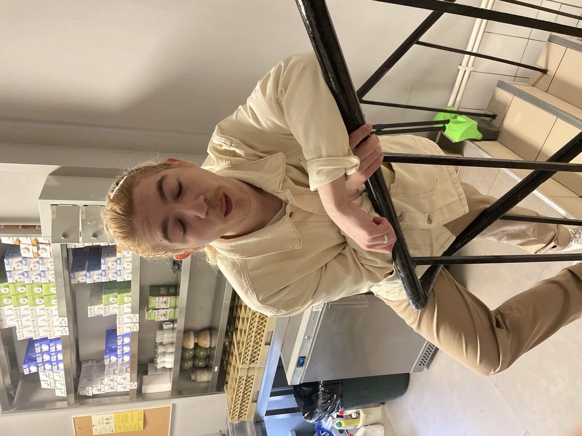 A young adult is holding onto a black metal railing while positioned on a staircase. The background suggests a commercial setting with shelving displaying packaged goods and a stainless steel counter. The location is not discernible as Moscow, Russia based on visual evidence.