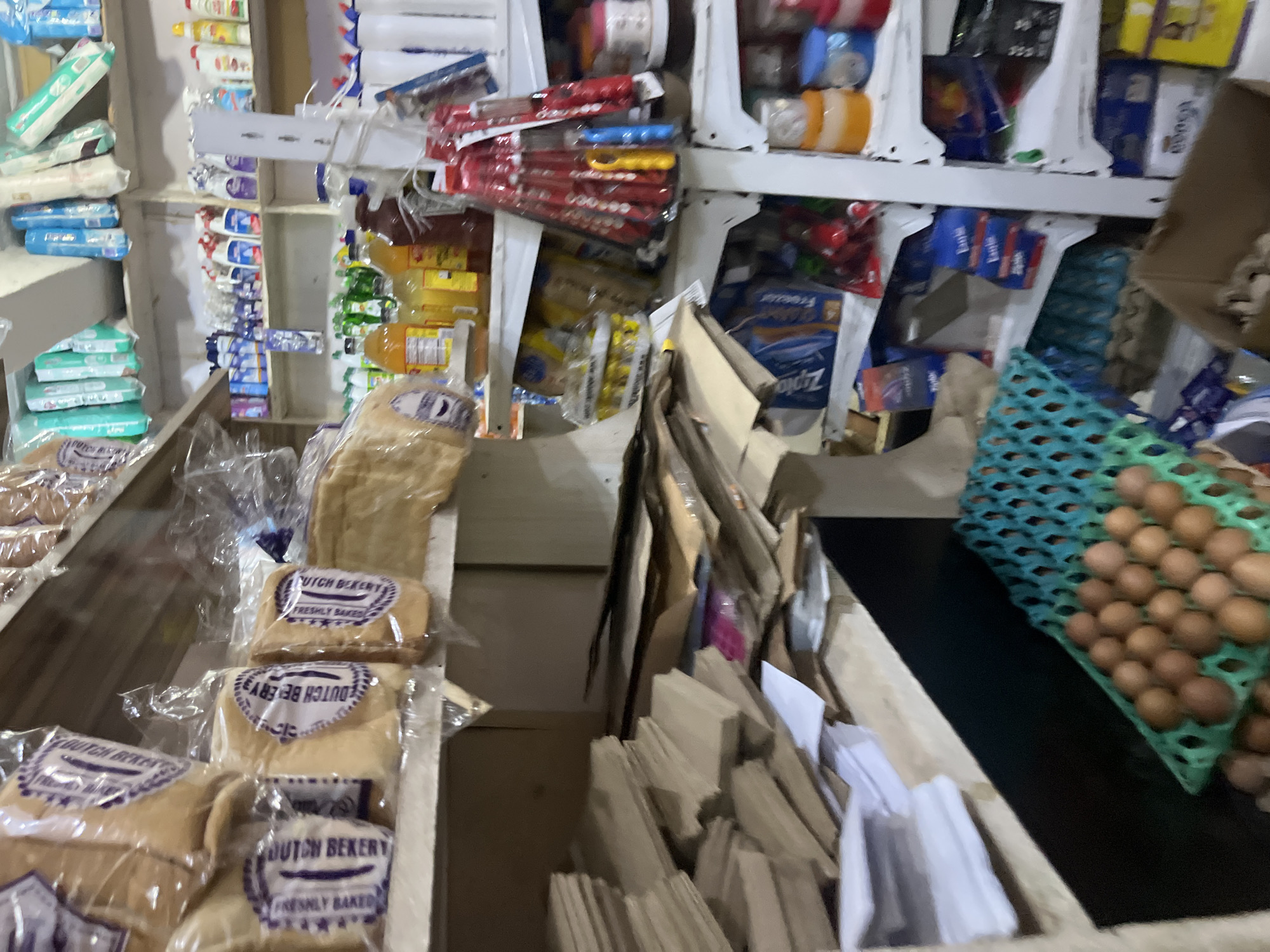 This is a medium shot image of a crowded Nigerian convenience store. The store is filled with a variety of goods, including packaged bread, bottled beverages, cleaning supplies, and food items. In the foreground, a green egg carton is filled with brown eggs. To the left, stacks of brown paper bags and white paper are piled on a shelf. In the background, shelves are stocked with various products. The lighting is dim, suggesting an indoor setting. The overall impression is one of a busy, well-stocked store in Bukuru, Nigeria.