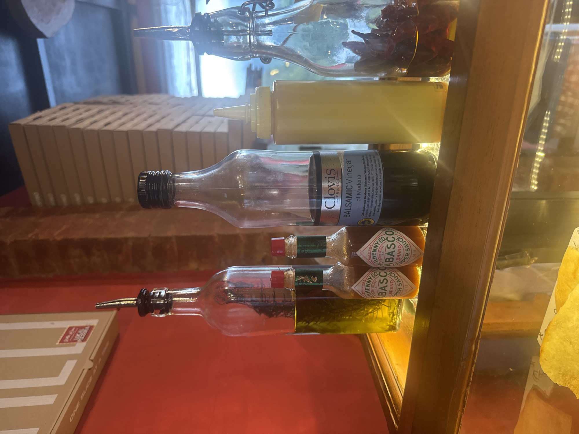The image depicts a shelf of condiments, likely in a restaurant or food service establishment. There are five bottles, each containing a different sauce or oil. From top to bottom, these are a clear oil with red peppers, a bottle of yellow mustard, a bottle of "Clovis" Balsamic Vinegar of Modena (imported by PT. Classic Fine Food Indonesia, Jakarta 14240 - according to the label), and two bottles of Tabasco sauce (60 ml each). The bottles appear to be displayed on a glass shelf within a wooden frame. A box from "One Slice at a Time" Local Pizzeria is visible on a red surface below, and there are potato chips visible on a glass shelf to the right. The location is Baturiti, Indonesia based on the Indonesian importer information for the Balsamic Vinegar.