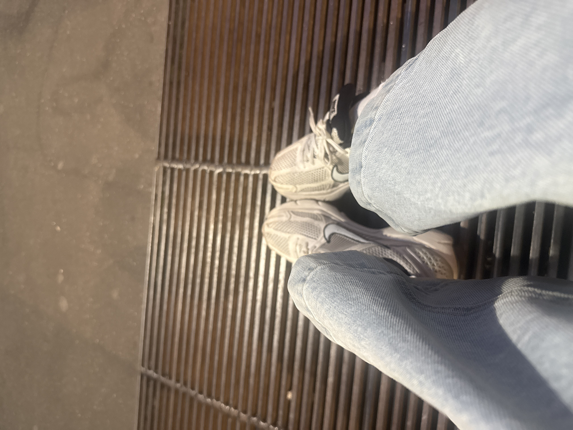 The image shows a person's-eye-view of their feet standing on a metal grate in Milan, Italy. The person is wearing light blue jeans and white Nike sneakers. The grate is made of evenly spaced metal bars. To the left of the grate is a solid concrete surface. The perspective suggests the person is standing still, looking down at their feet. The lighting indicates it is likely daytime, and there are no other people or objects visible in the immediate vicinity. The location is characterized by urban infrastructure, suggesting a public space or pedestrian area in Milan.