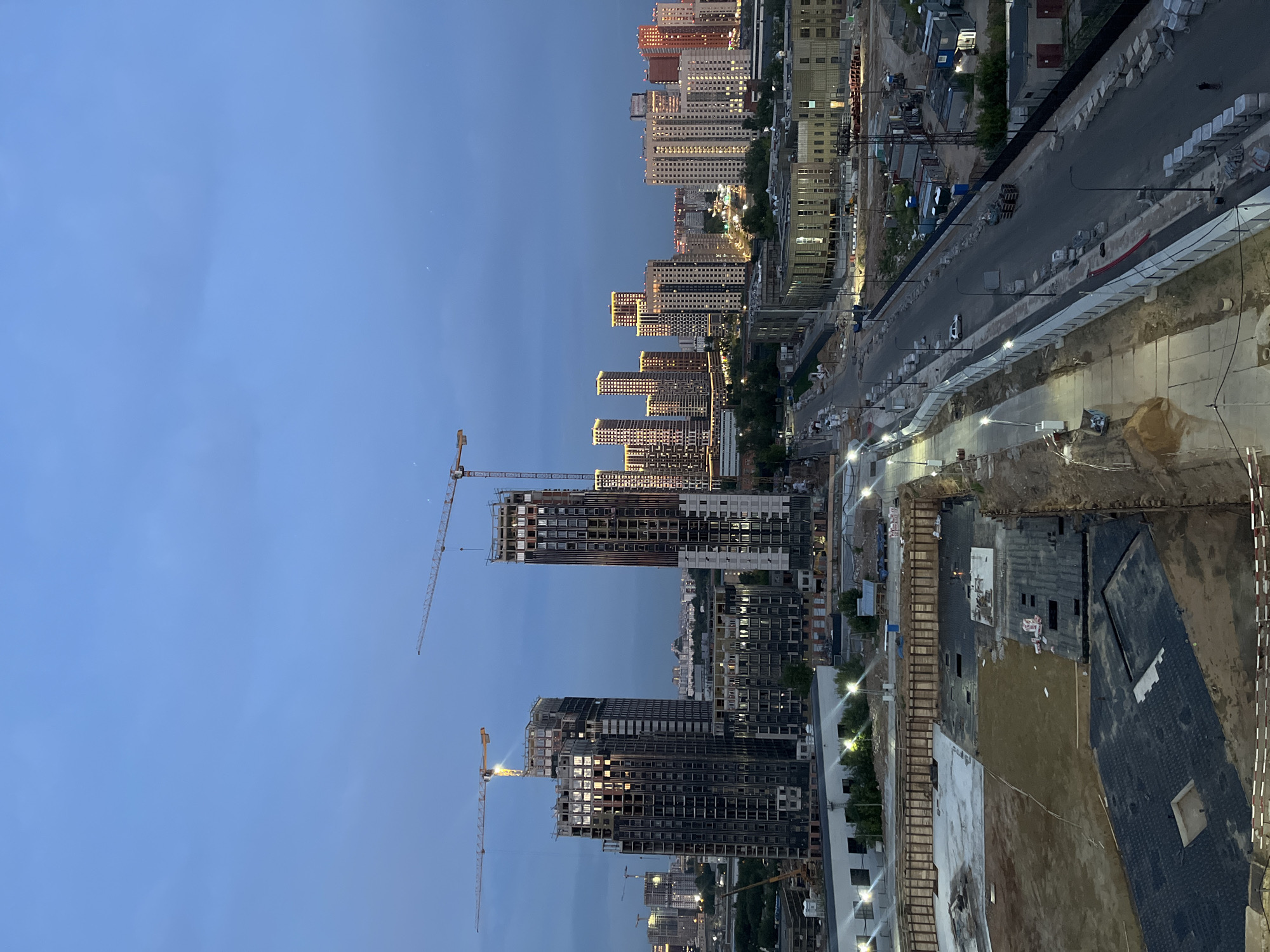 The image depicts an urban construction site in Tarasovka, Russia, during twilight. Several high-rise buildings are under construction, with cranes towering over them. The foreground shows an excavated area with various construction materials, including large black mats laid out and sand. A paved road with streetlights runs alongside the construction site, with a few cars visible. In the background, completed residential buildings are illuminated, creating a stark contrast with the unfinished structures. The sky is a deep blue, indicating the late evening hour, with faint clouds present. No people are clearly discernible in the image, and the overall scene is one of ongoing development and urban expansion.