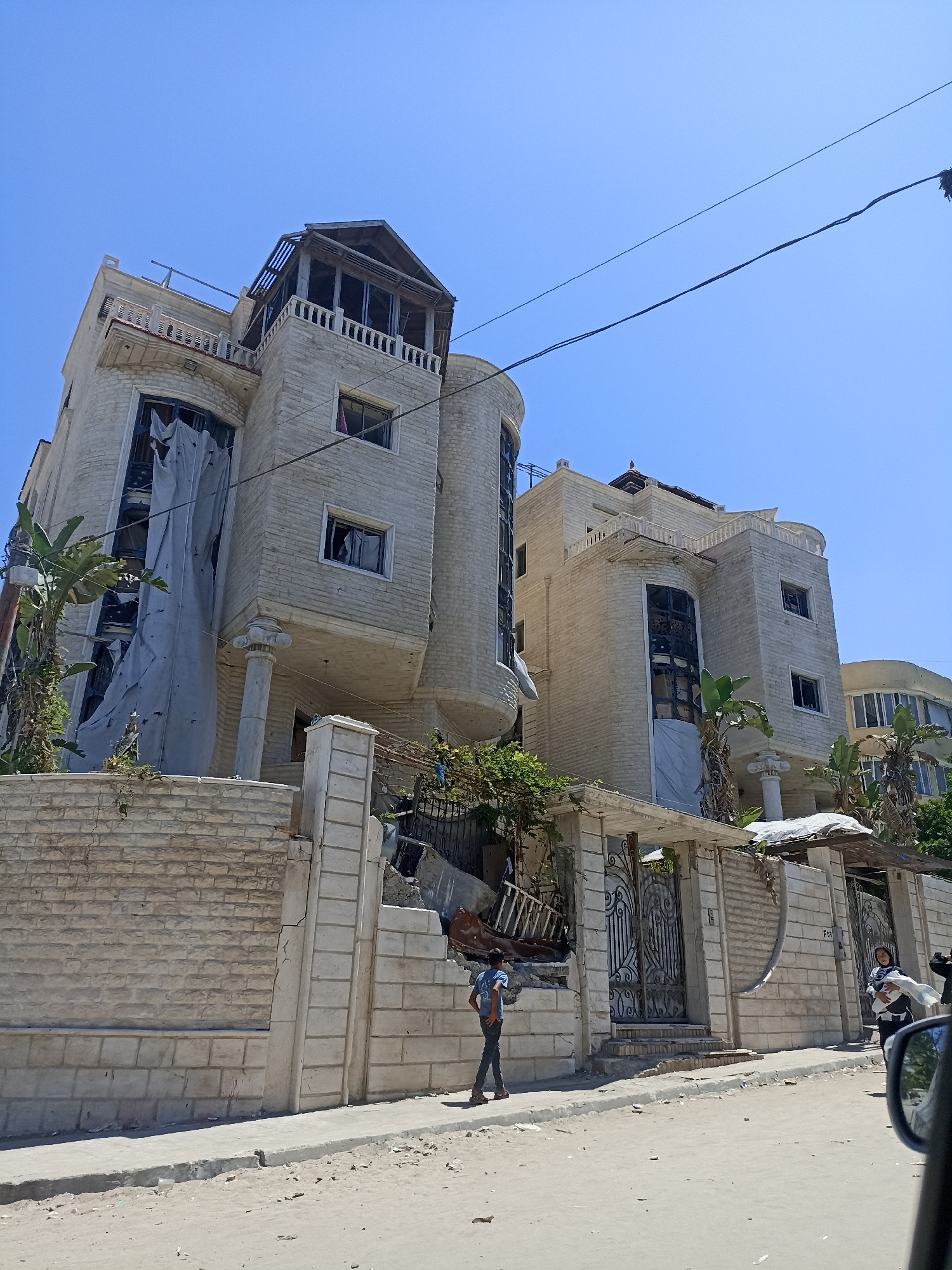 The image shows a three-story building in Gaza City, Palestinian Territories. The building appears to be made of light-colored stone and has a modern architectural style with curved sections and balconies. There is evidence of damage to the building, including broken windows and what appears to be debris around the lower floors. A large section of fabric is hanging down the side of the building, covering part of a window. A stone wall with metal gates surrounds the property. Two individuals are visible on the sidewalk in front of the building. One is a young male wearing a blue t-shirt and dark pants, standing near the gates. Another individual, a female, is walking towards the gates carrying a white bundle. The street in front of the building is paved with concrete and appears to be sandy. The sky is clear and blue.