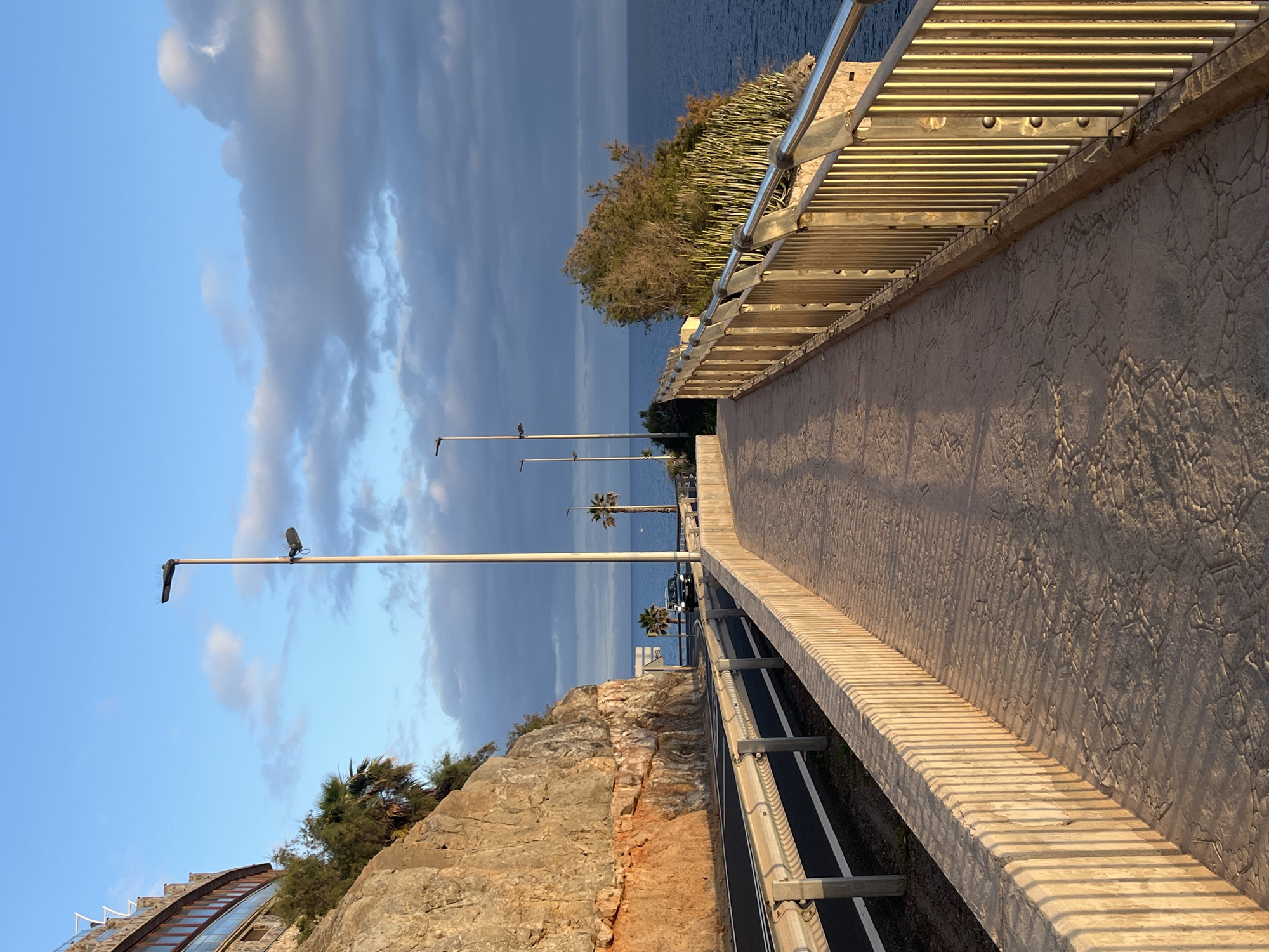 The image shows a promenade or bridge-like structure overlooking the sea in Puerto Rico, Spain. The structure is constructed with a stone-like walkway and features metal railings along both sides. Adjacent to the walkway, on one side, there's a road with a barrier. Street lamps are visible along the roadside. The landscape includes rocky cliffs and vegetation. The sky is a mix of blue and gray clouds. In the distance, palm trees are visible near the water. There are no people visible in the image.