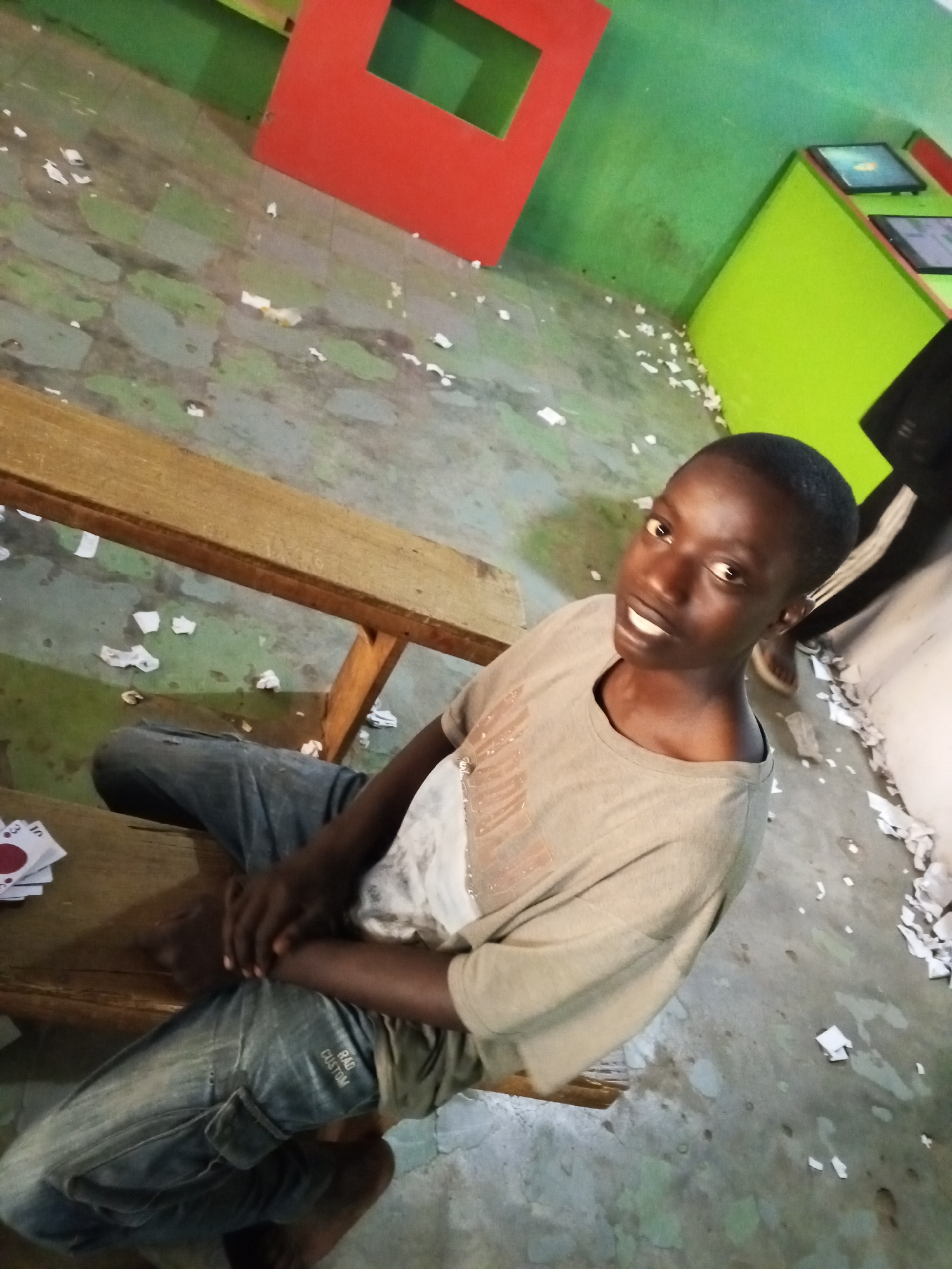 A young, dark-skinned person with short hair sits on a wooden bench in an interior space. They are wearing a light-colored t-shirt and blue jeans. The floor is littered with white scraps. The room is painted in green, red, and grey. Two computer screens are visible on a green cabinet. A partial view of another person's legs is visible in the background. This photo was taken in Gwong, Nigeria.