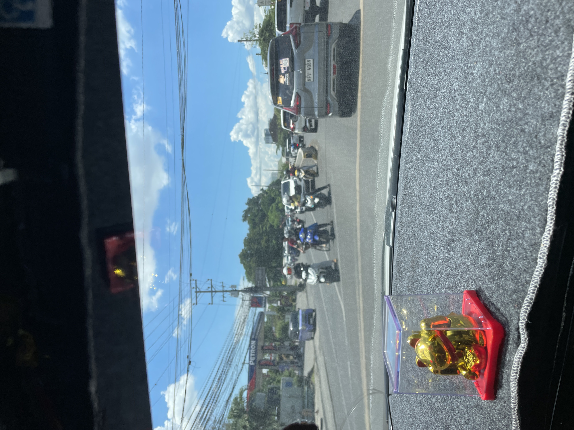 The image depicts a street scene in Davao City, Philippines, viewed from inside a vehicle. The foreground features a dashboard with a small, gold, lucky cat figurine in a clear plastic case. Beyond the dashboard, the street is visible with multiple vehicles, including cars and motorcycles, in traffic. A Petron gas station is identifiable on the left side of the road, with trees and utility poles lining the street. The sky is blue with scattered clouds, suggesting a sunny day.