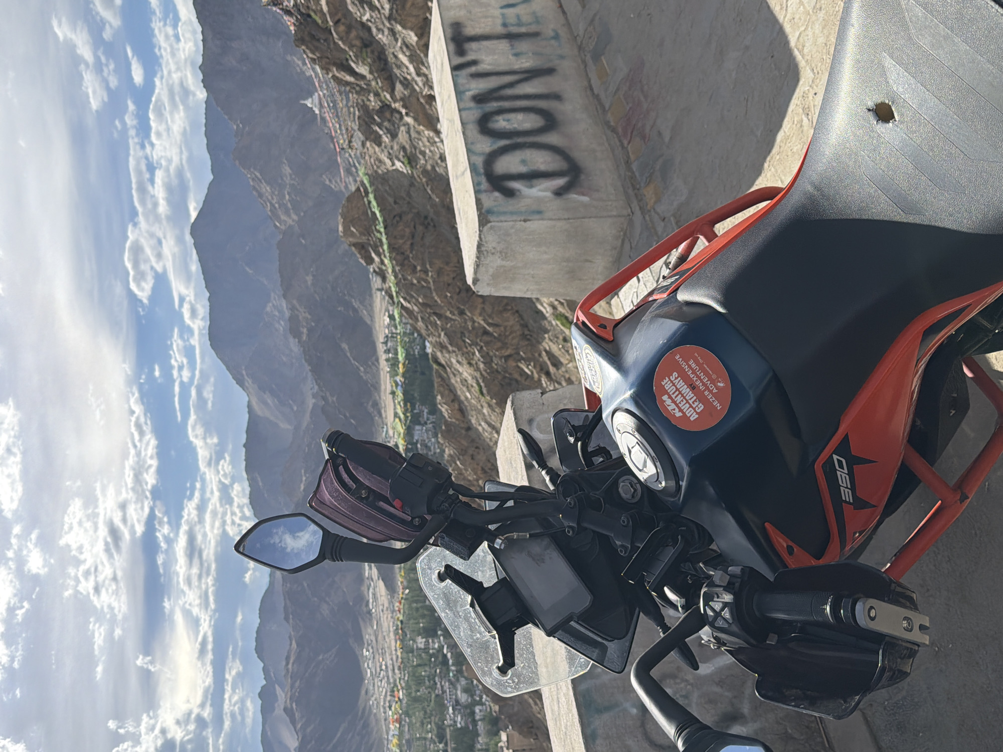 A motorcycle is parked in front of a mountainous landscape. The motorcycle is a KTM 390 Adventure, with a red frame and blue and orange body. A sticker on the fuel tank reads "KTM ADVENTURE GETAWAYS BY NEZER INEXPENSIVE ADVENTURE". The motorcycle is parked on a concrete barrier with graffiti that says "DON'T". In the background, there are mountains with a village nestled in the valley below. The sky is partly cloudy. This scene is consistent with the Shanti Stupa viewpoint in Leh, India, offering panoramic views of the Himalayas and the town.