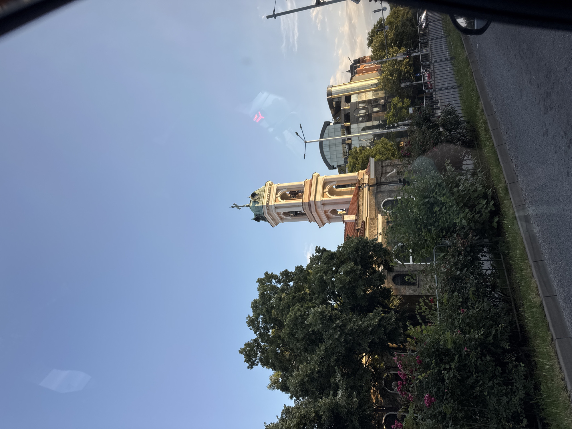 The image presents a street-level view in the city of Plovdiv, Bulgaria, captured from within a moving vehicle during what appears to be late afternoon or early evening.
The central and most prominent feature is the Catholic Cathedral of St. Ludwig (St. Louis), identifiable by its distinctive bell tower. The tower is constructed from light-colored stone or stucco, featuring arched bell openings, a green domed roof, and a cross at its apex.
To the left of the cathedral, a dense cluster of mature green deciduous trees partially obscures an older stone building with arched windows. In the immediate foreground on the left, a metal barrier runs parallel to the road, behind which are planted dense green bushes, some bearing pink blossoms. The ground between the barrier and the road is grass.
To the right of the cathedral, a series of contemporary multi-story buildings line the street. One building exhibits a reflective glass facade and a partially visible sign that reads "VICTORIA." Further down the street, additional buildings with varied architectural styles and color palettes are apparent.
The sky overhead is predominantly clear and light blue, with a few wispy white clouds visible on the right side. Reflections are prominent in the upper portion of the image, including a distinct bright pink, stylized "Puma" logo in the upper right quadrant and a rectangular light reflection in the upper left.
The photograph's perspective is from a vehicle, indicated by the dark frame of a car window and a partial view of a side-view mirror on the far right. The foreground consists of a paved asphalt road, which stretches into the distance. Distant traffic is visible on the road, including a red car and a white car. Streetlights are positioned along the roadway. No individuals are distinctly visible or engaged in specific actions within the frame.