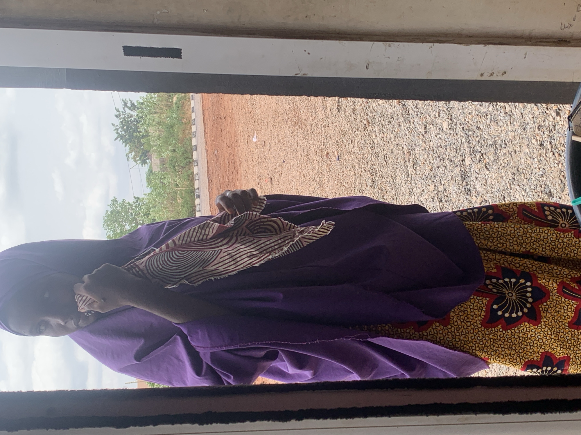 A medium shot depicts a woman with dark skin, clad in a purple robe and head covering, standing outdoors, viewed from a partially obscured vantage point behind a white frame. She's holding a brown and white patterned cloth in her hand, raising it to her face, potentially wiping it. A patterned yellow and brown garment is visible under the purple robe. Outside the frame, a sandy ground is visible, leading to a tree line and sky, indicating a location within the city of Adankolo, Nigeria.