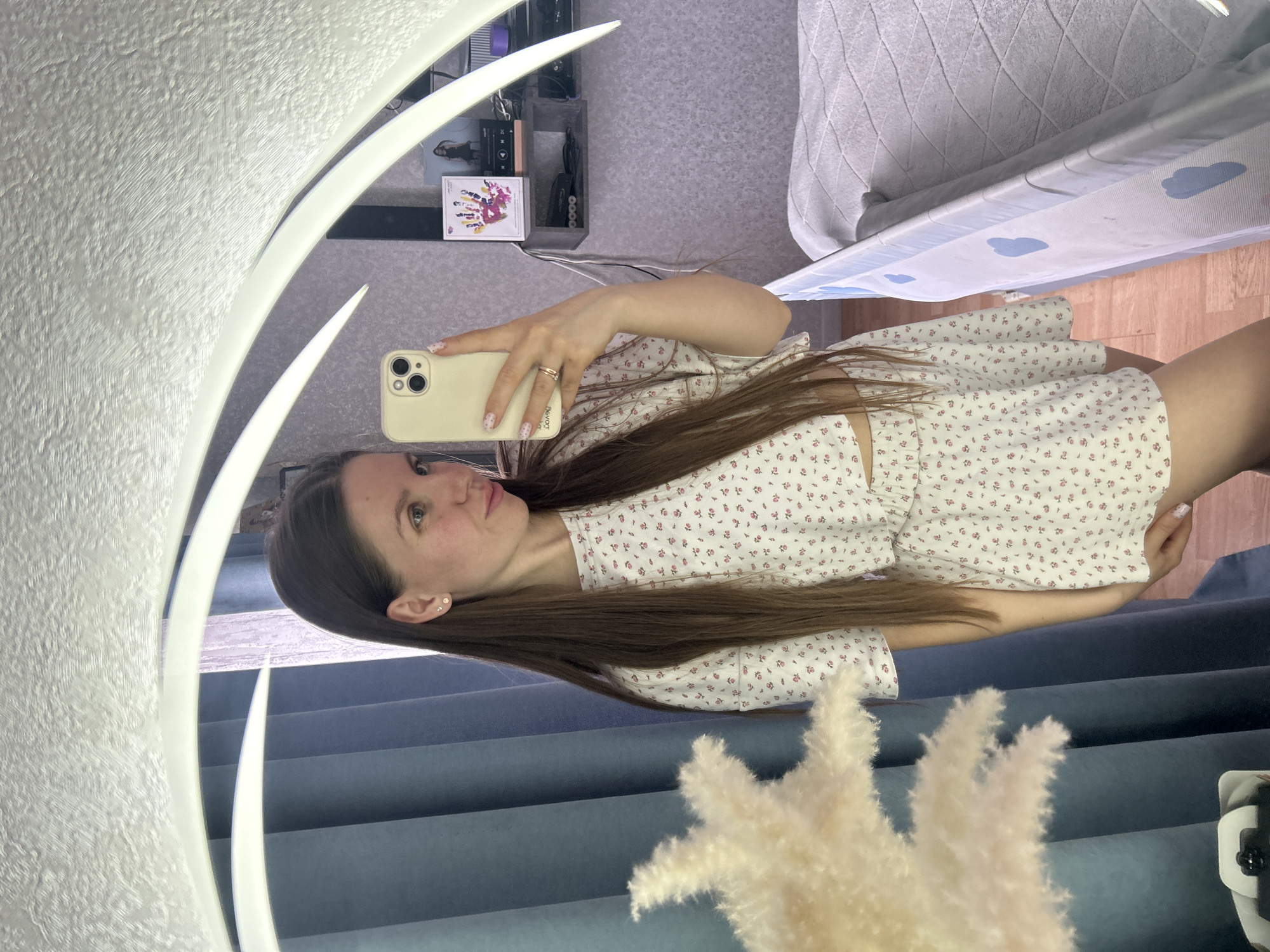 A woman takes a selfie in a mirror. She is wearing a floral print outfit. The background includes a curtain, a bed, and a shelf with various items, including what appears to be a framed picture. The city of Klin, Russia, is not evident in the image.