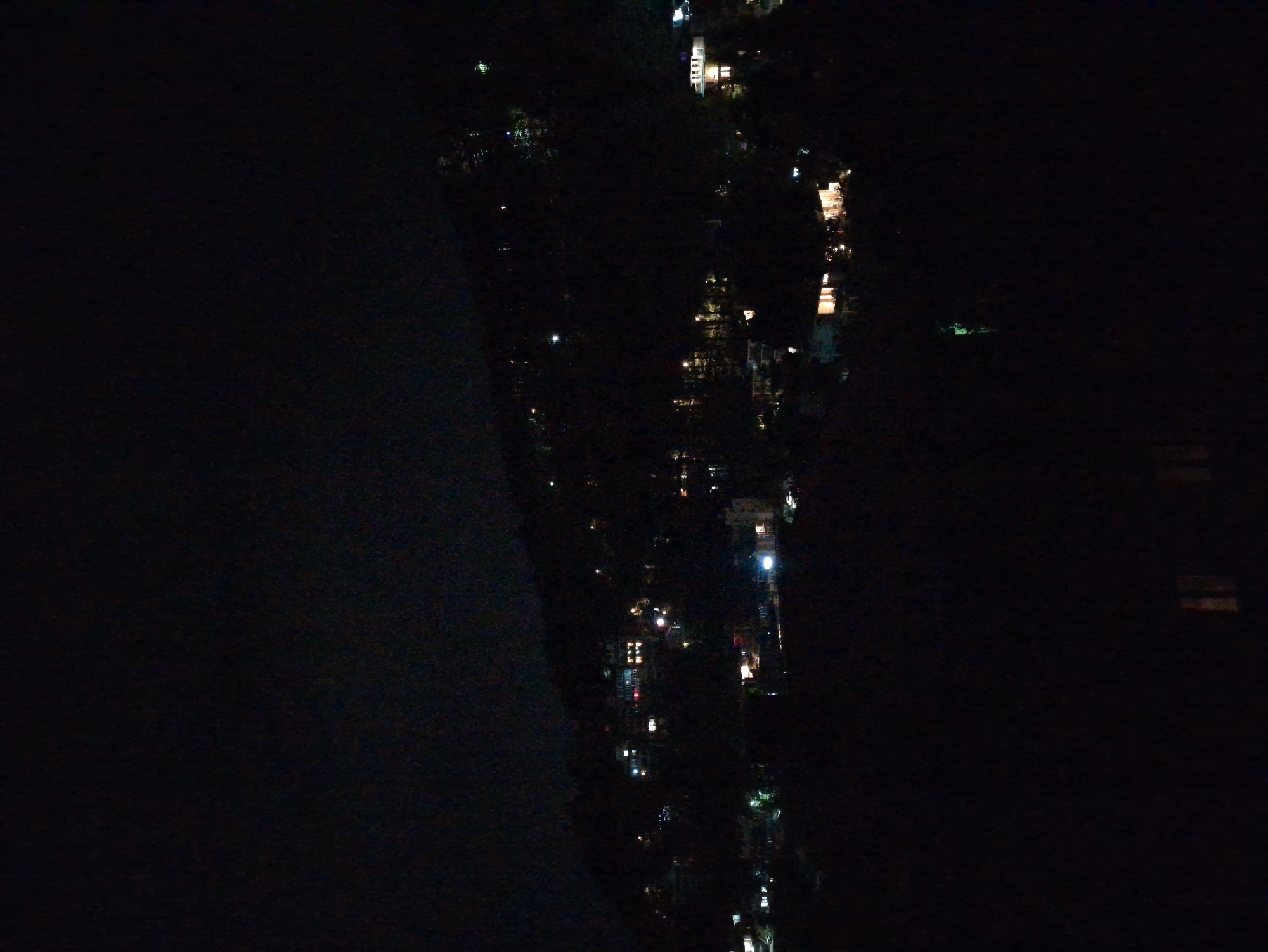The image depicts a nighttime view of a hilly landscape illuminated by scattered artificial lights from buildings. The lights suggest a settlement or town on the slopes of the hill. The darkness of the night obscures most details of the terrain and structures, with the exception of the glowing windows and lights of the buildings. There is no visible sign of people, specific activities, or interactions. The environment appears to be natural, with the human presence indicated by the presence of constructed buildings and lighting. The image does not contain enough visual information to identify the specific location within Haini, India.