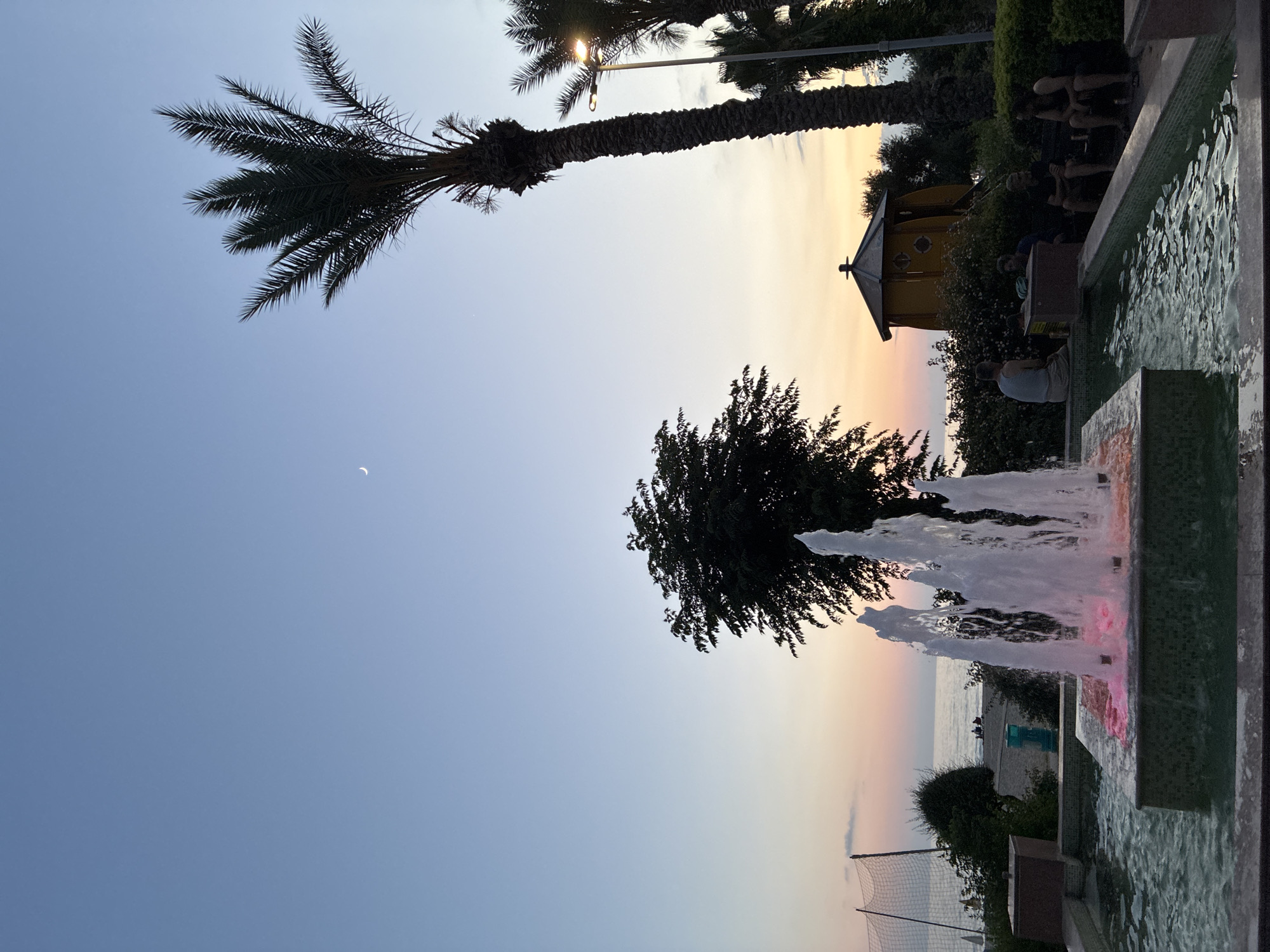 This is a vertical, eye-level, medium shot of a fountain with water jets illuminated by pink and white lights. The fountain's basin is tiled with green and blue mosaic tiles. In the background, a palm tree extends across the sky, its fronds silhouetted against a clear blue sky with a crescent moon visible. The horizon features the sea at sunset, with soft pastel colors in the sky. A yellow, octagonal gazebo or playhouse is visible behind some trees. Several people are seated on benches in the background, with one person sitting by the fountain. The scene evokes a tranquil evening ambiance. It is likely dusk or early evening in Alanya, Turkey, given the presence of palm trees and the sea.