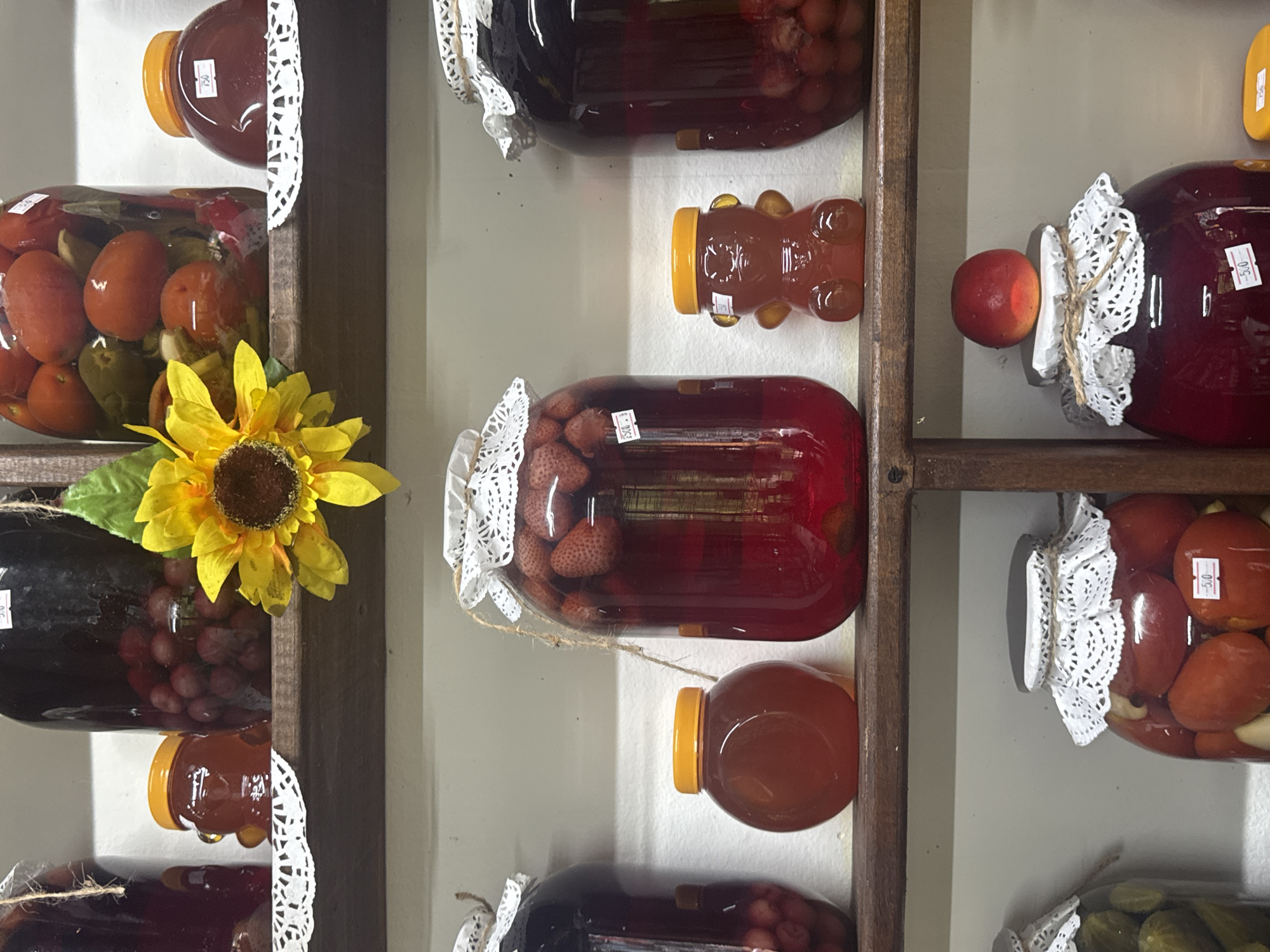 In Semenovka, Russia, a collection of preserved goods is displayed on wooden shelves. The top shelf holds several large glass jars, one filled with red grapes, another with what appear to be tomatoes and green peppers, and a third with preserved fruit in a dark liquid. A decorative sunflower is placed prominently on this shelf. The middle shelf features three jars of preserves: a large jar of strawberries in a red liquid, a smaller round jar of honey, and a bear-shaped jar, also filled with honey. The bottom shelf contains more jars of preserves, including pickles, tomatoes, and a large jar with a single red fruit on top, all covered with decorative fabric and tied with twine.