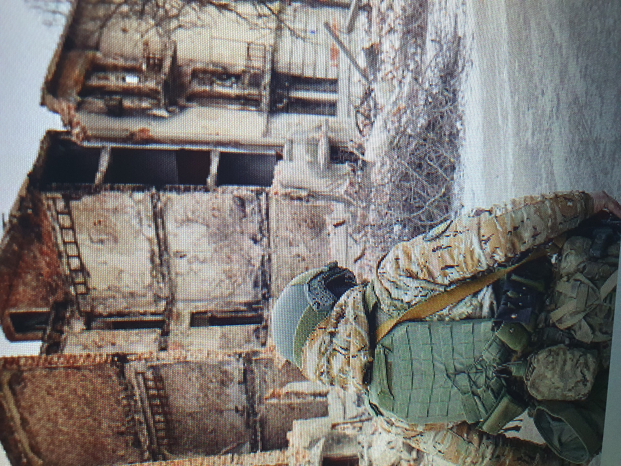 The image depicts a person in military camouflage and protective gear, facing away from the viewer. The person is wearing a helmet, a tactical vest with pouches, and appears to be equipped with a backpack. They are standing in what looks like a war-torn urban environment. The background shows a severely damaged brick building with exposed structural elements and rubble scattered around. The building's facade is crumbling, with gaping holes where windows and doors once were. The debris in the foreground suggests destruction and decay. The scene is set against a muted, overcast sky. There is no indication of the specific location being Tashkent, Uzbekistan.