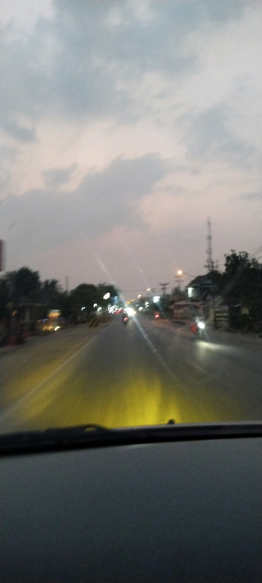 The image depicts a view from inside a vehicle, looking out at a road in Kali Balangan, Indonesia, during twilight. The sky is overcast with gray and light pink clouds. The road is paved and appears to be a two-lane highway with streetlights and lights from buildings lining the sides. Several vehicles, including motorcycles with illuminated headlights, are visible on the road, suggesting moderate traffic. The headlights of oncoming vehicles cast strong yellow light onto the road. Trees and structures, possibly houses or shops, are visible along the roadside. A cellular tower is present in the distance. The foreground shows the dashboard and windshield of the vehicle, with a blurred yellow glow emanating from the headlights.