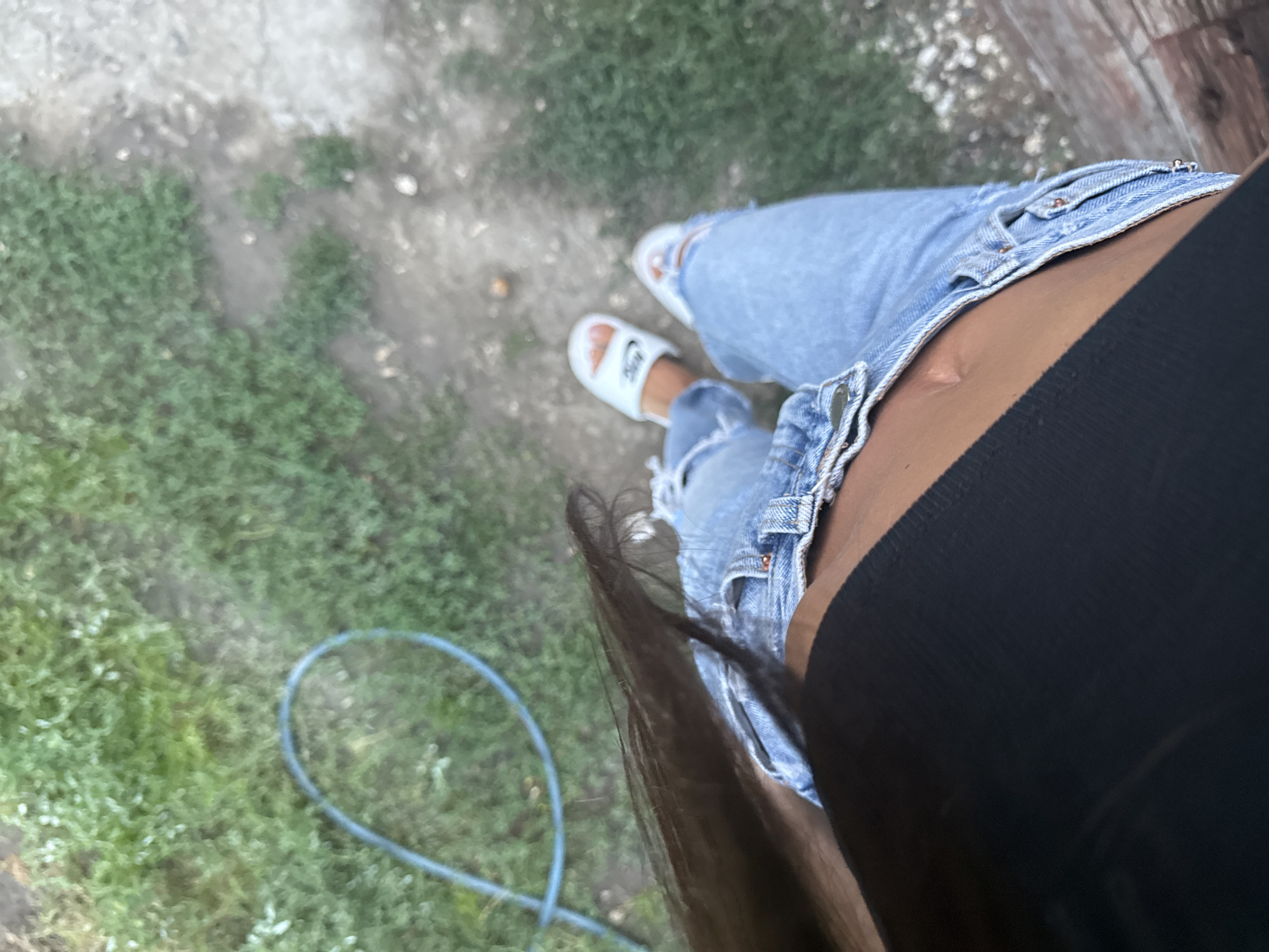 A first-person perspective shot shows a person from the waist down, wearing ripped light-wash denim jeans and white slide sandals. The individual is standing on a dirt and grass ground, with a blue hose looped on the ground to the left. The background is blurred, but appears to be an outdoor setting with vegetation and a wooden structure. The lighting suggests it is daytime. The location of Basarabeasca, Moldova is not discernible from the image.